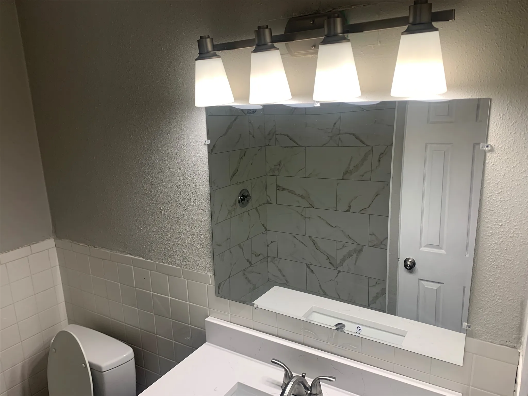 Bathroom featuring a four-light vanity fixture, a large wall-mounted mirror, a white integrated sink with a chrome faucet, and white subway tile wainscoting