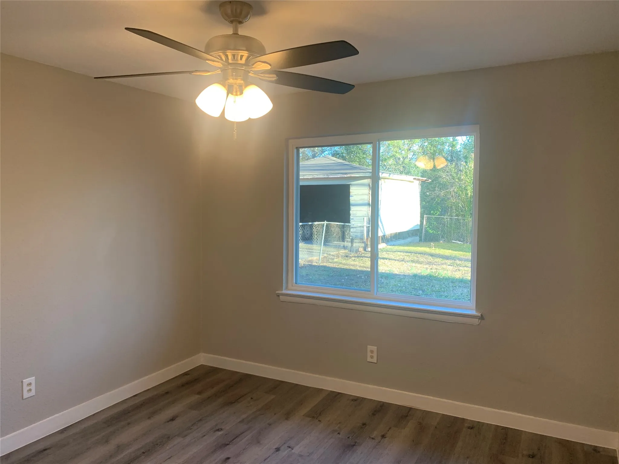 Room featuring a large window, neutral wall color, wood-finish flooring, white baseboards, and a ceiling fan with integrated lighting