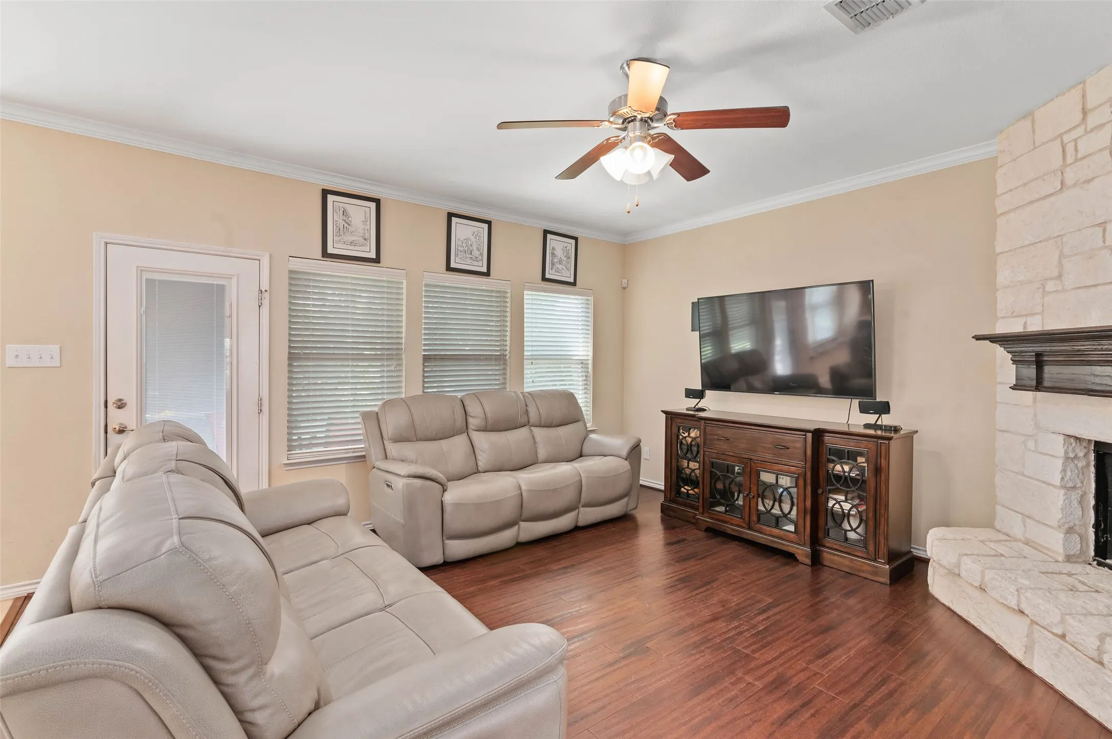 Warm living area featuring wood-finish flooring, a stacked stone fireplace with a dark wood mantel, and a ceiling fan with integrated lighting