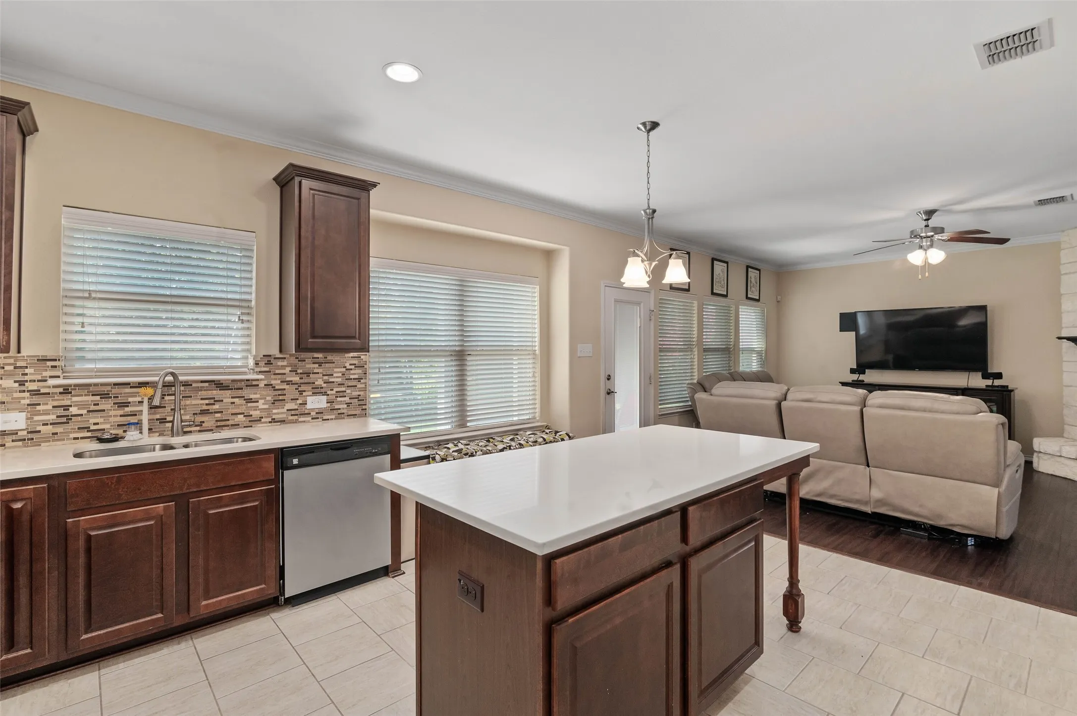 Open-concept kitchen and living area featuring a kitchen island with a light-toned countertop, wood-finish cabinetry, stainless steel dishwasher, mosaic tile backsplash, and a ceiling fan in the living space