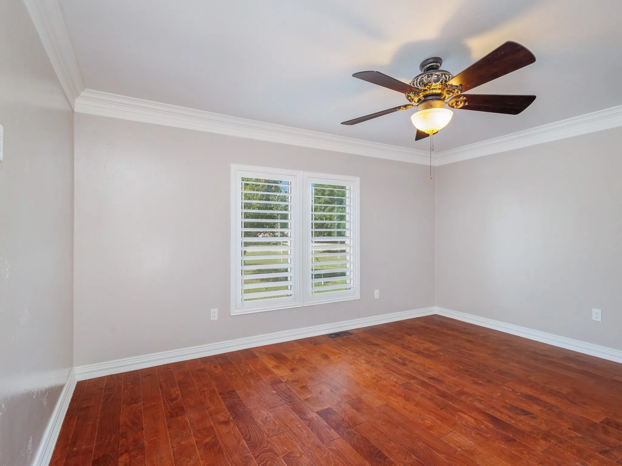 Spacious room featuring wood-finish flooring, two windows with white plantation shutters, crown molding, and a ceiling fan with integrated lighting