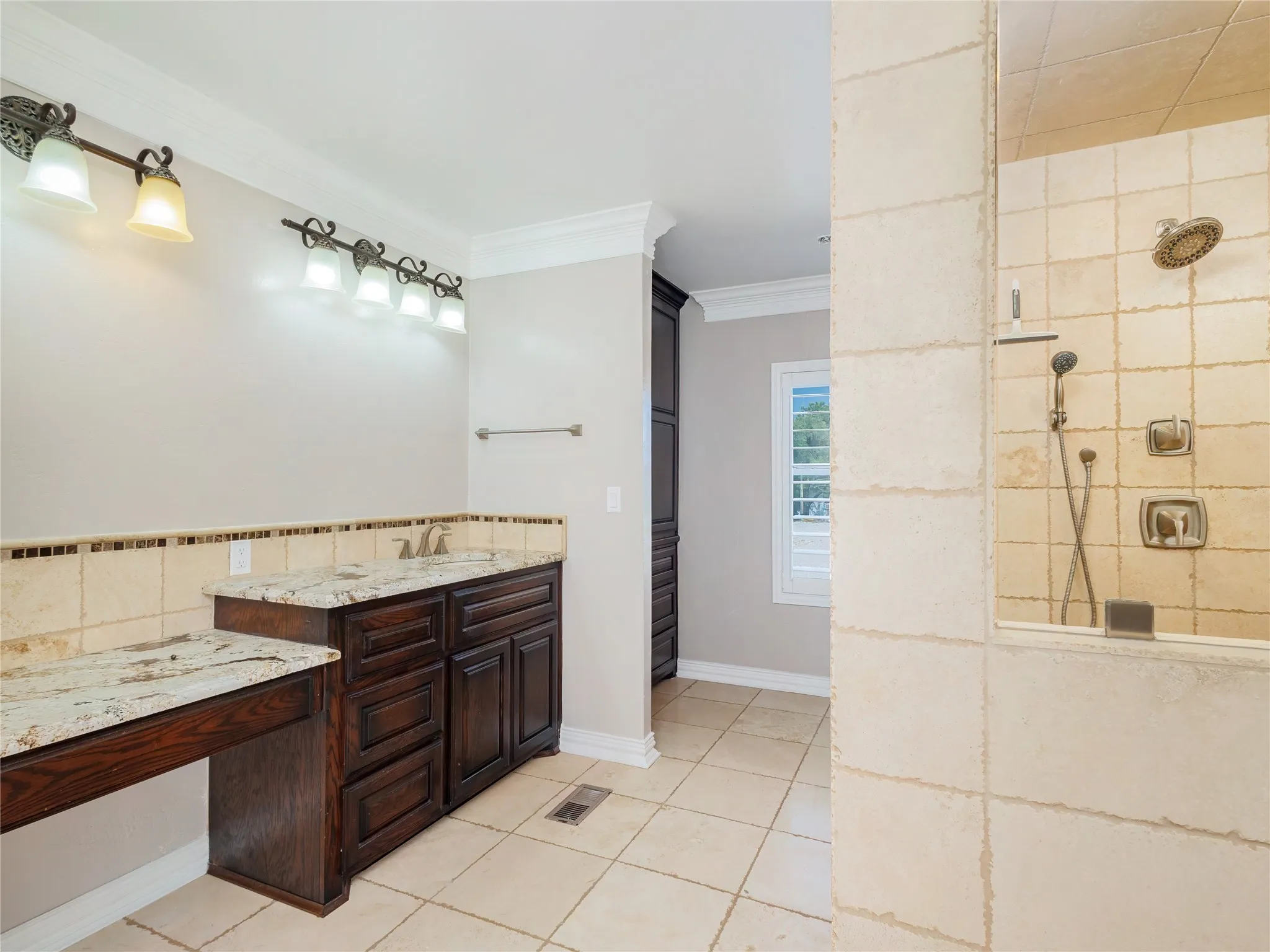 Expansive bathroom featuring a tiled walk-in shower with dual shower heads, a double vanity with granite countertops, rich wood cabinetry, and tiled flooring
