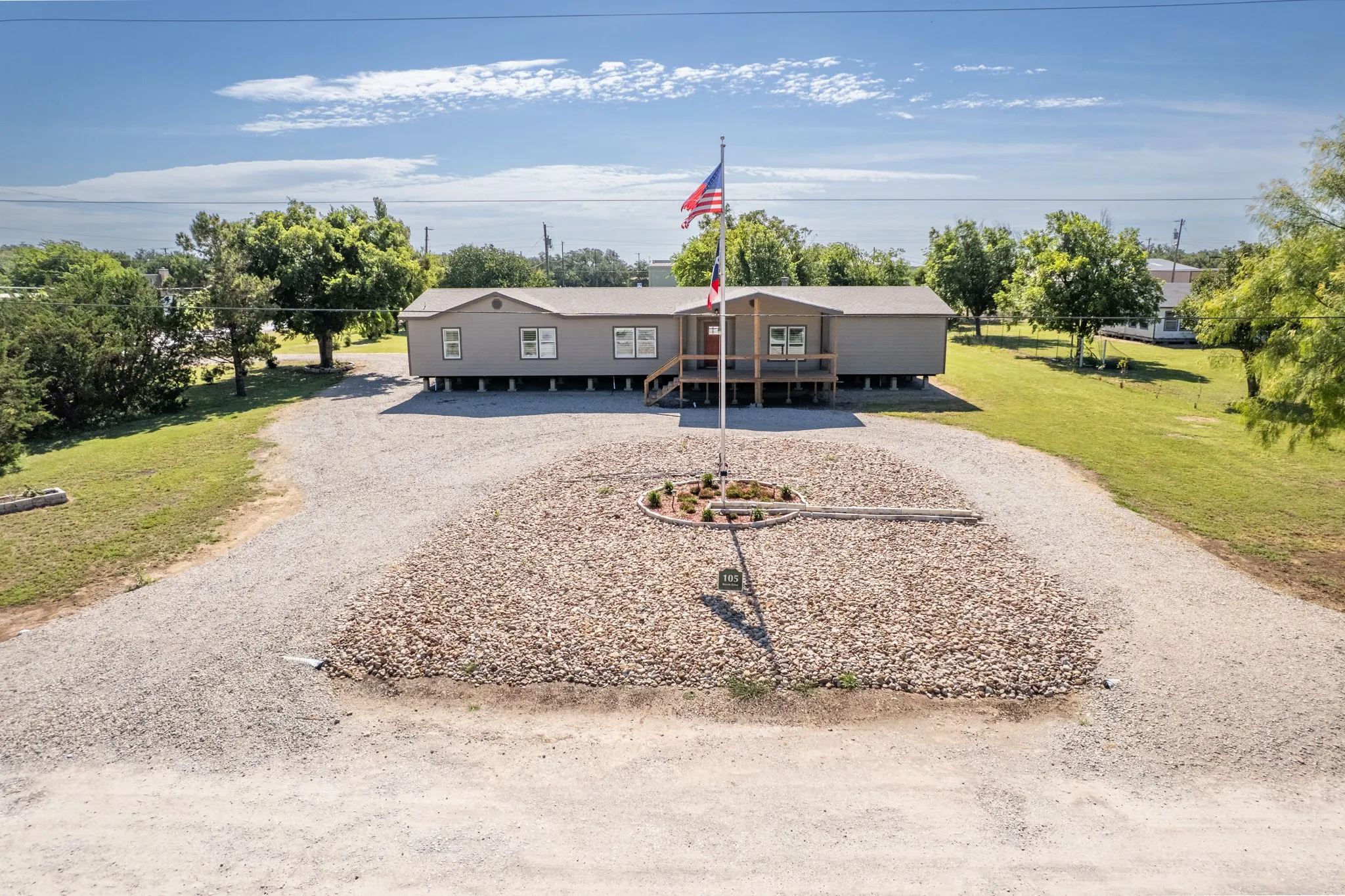 Elevated single-story residence featuring a covered front porch, gravel driveway, and a central rock garden with flagpole