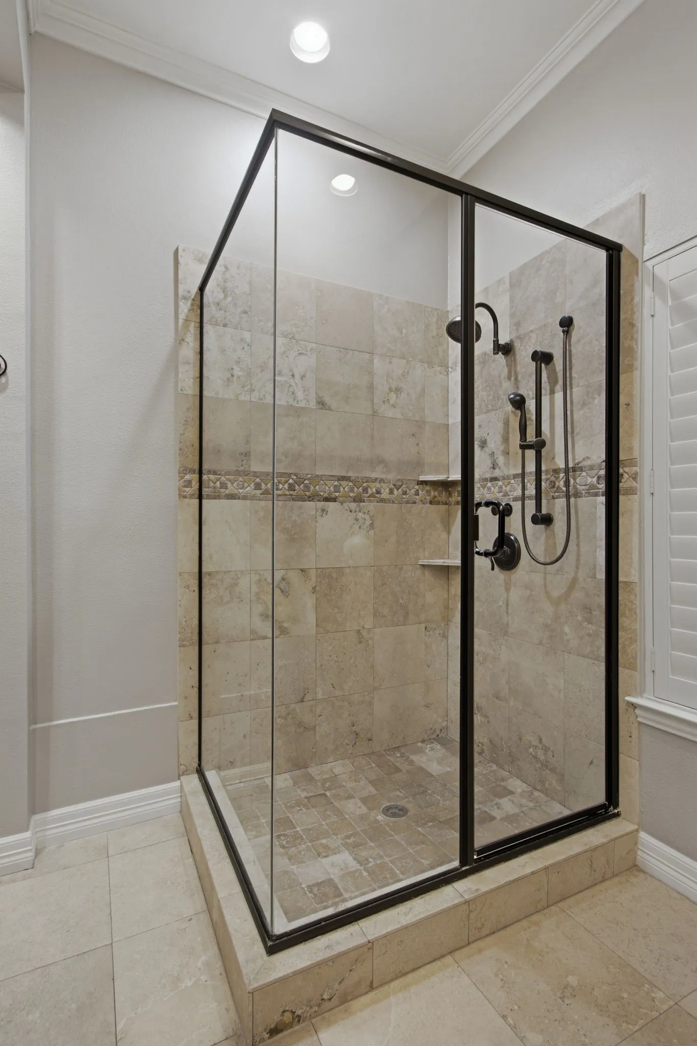 Walk-in shower featuring glass enclosure with dark bronze framing, full-height stone tile surround, integrated shower shelf, and coordinating dark bronze fixtures including a rain showerhead and handheld sprayer