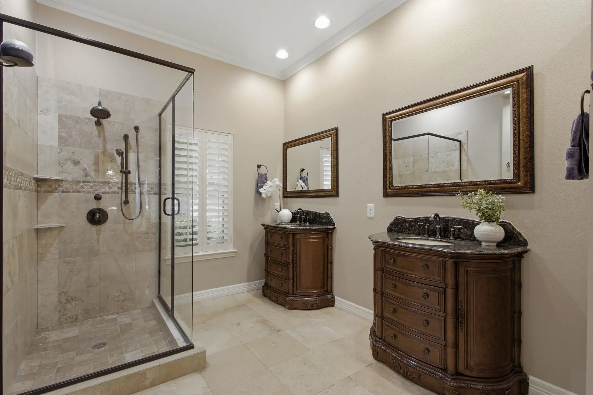 Spacious bathroom featuring a glass-enclosed shower with multiple showerheads, twin vanity units with dark stone countertops, decorative framed mirrors, and plantation shutters on the window