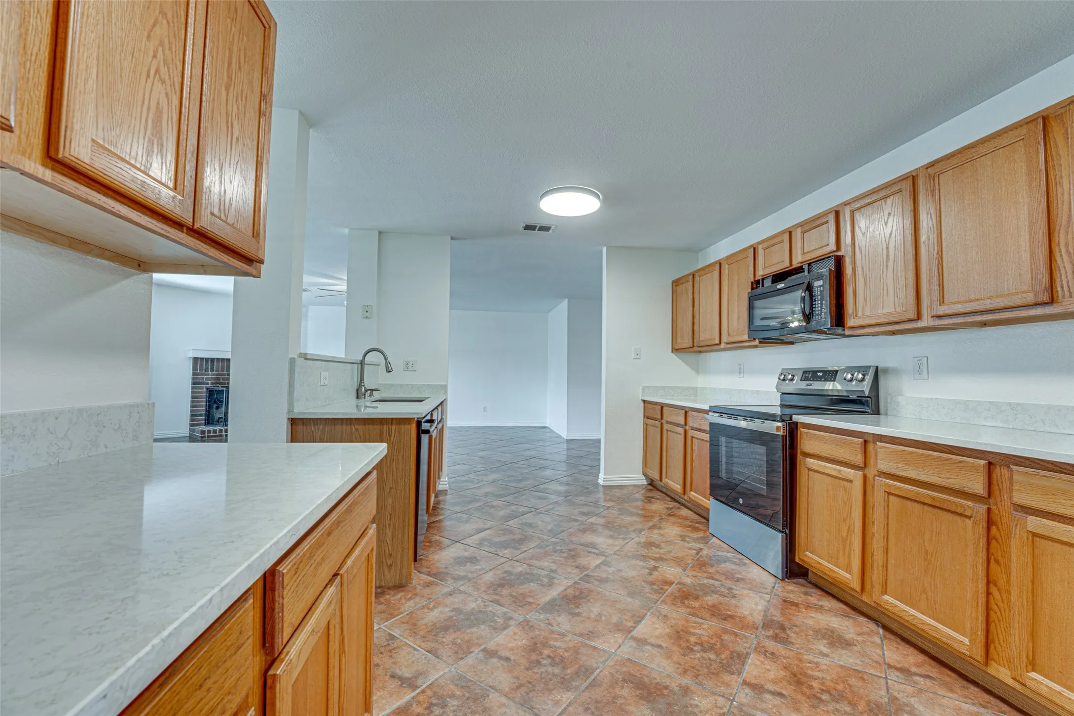 Kitchen featuring light wood cabinetry, light-toned countertops, a stainless steel sink with a chrome faucet, black appliances including a range and microwave, and brown-toned tile flooring