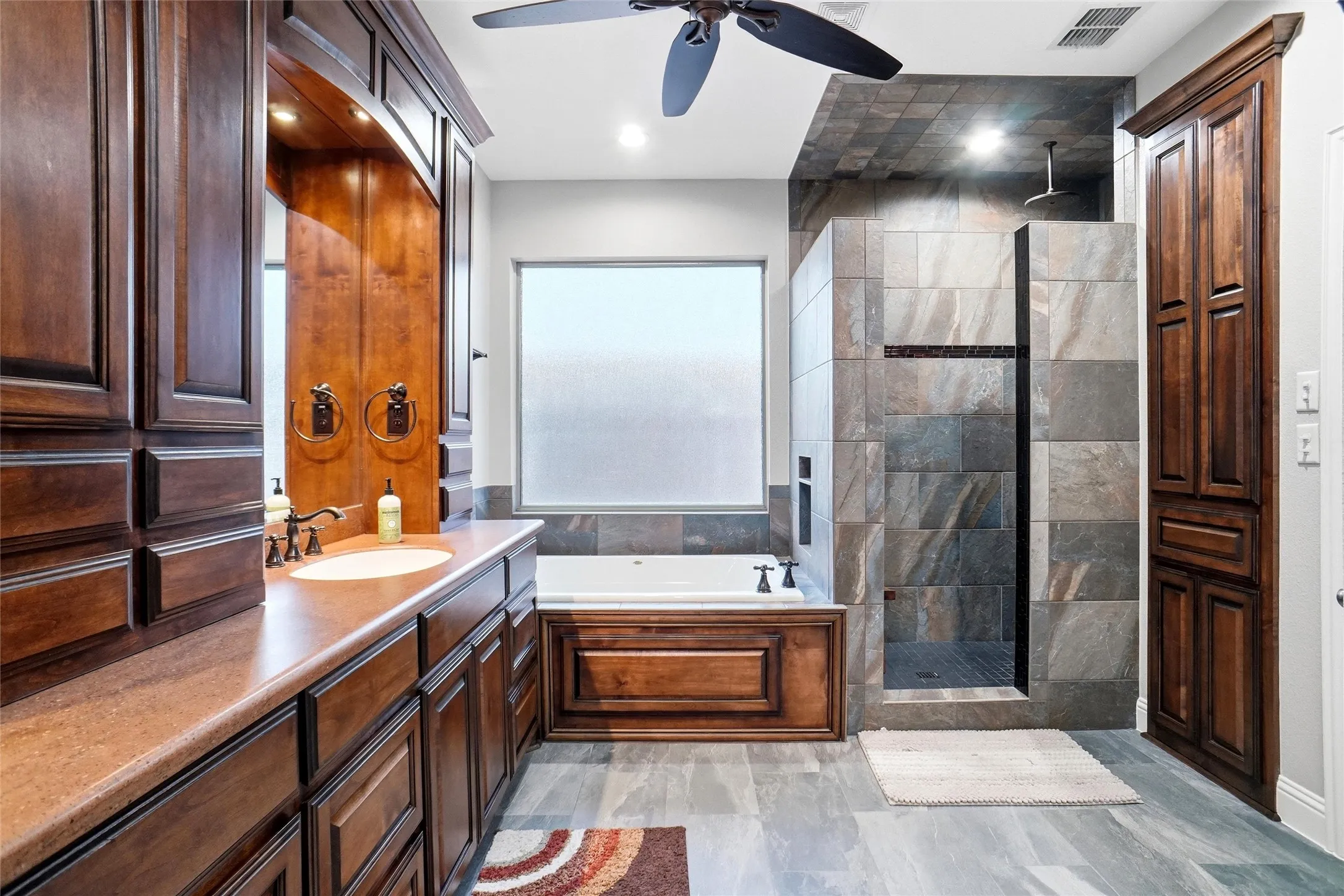 Bathroom featuring a built-in soaking tub with wood-finish paneling, a walk-in shower with tile surround, a built-in vanity with integrated sink and storage cabinetry, a ceiling fan, and overhead lighting