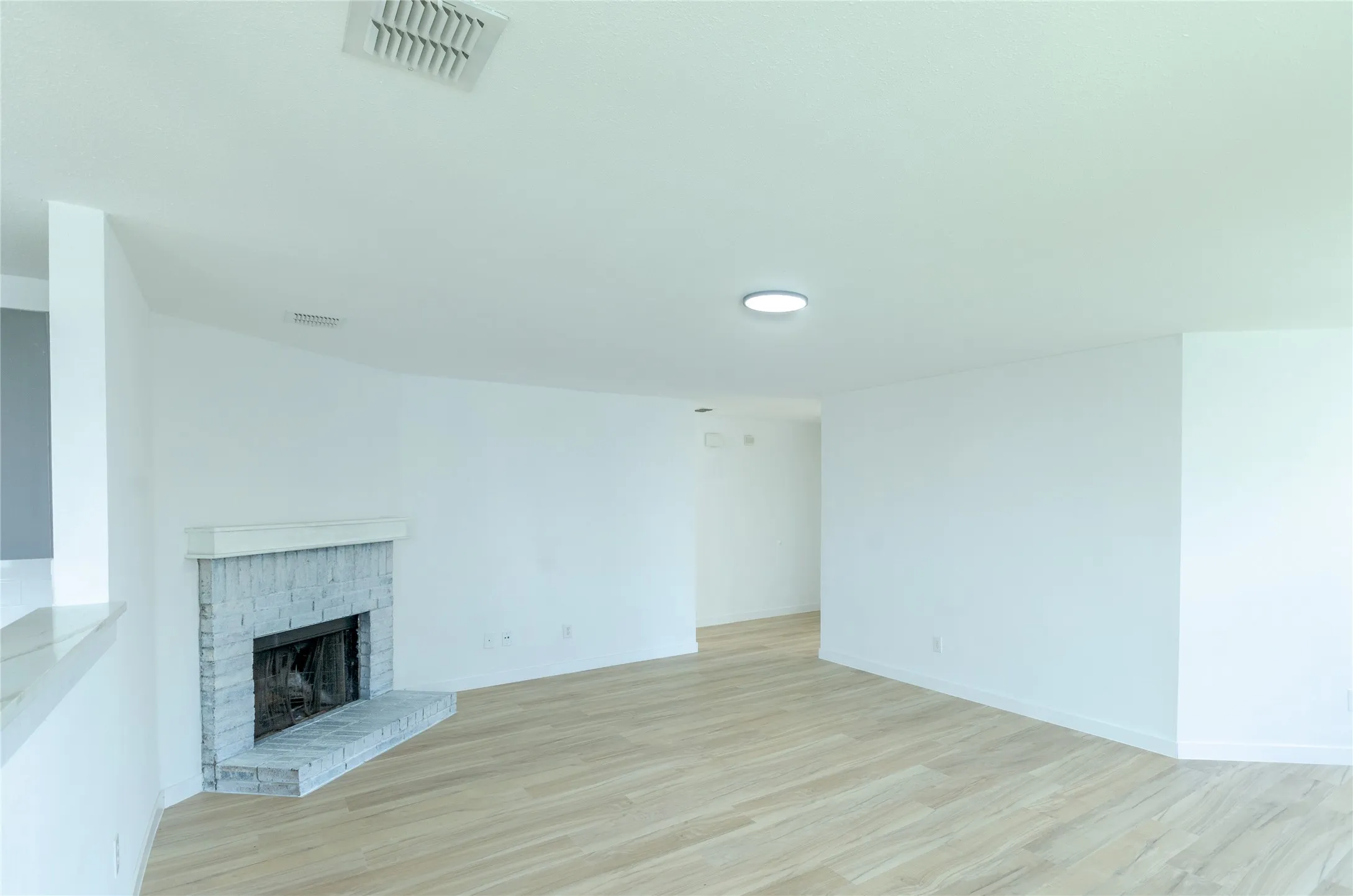 Spacious room featuring light wood-finish flooring, a white brick fireplace with a stone mantel, and recessed ceiling lighting