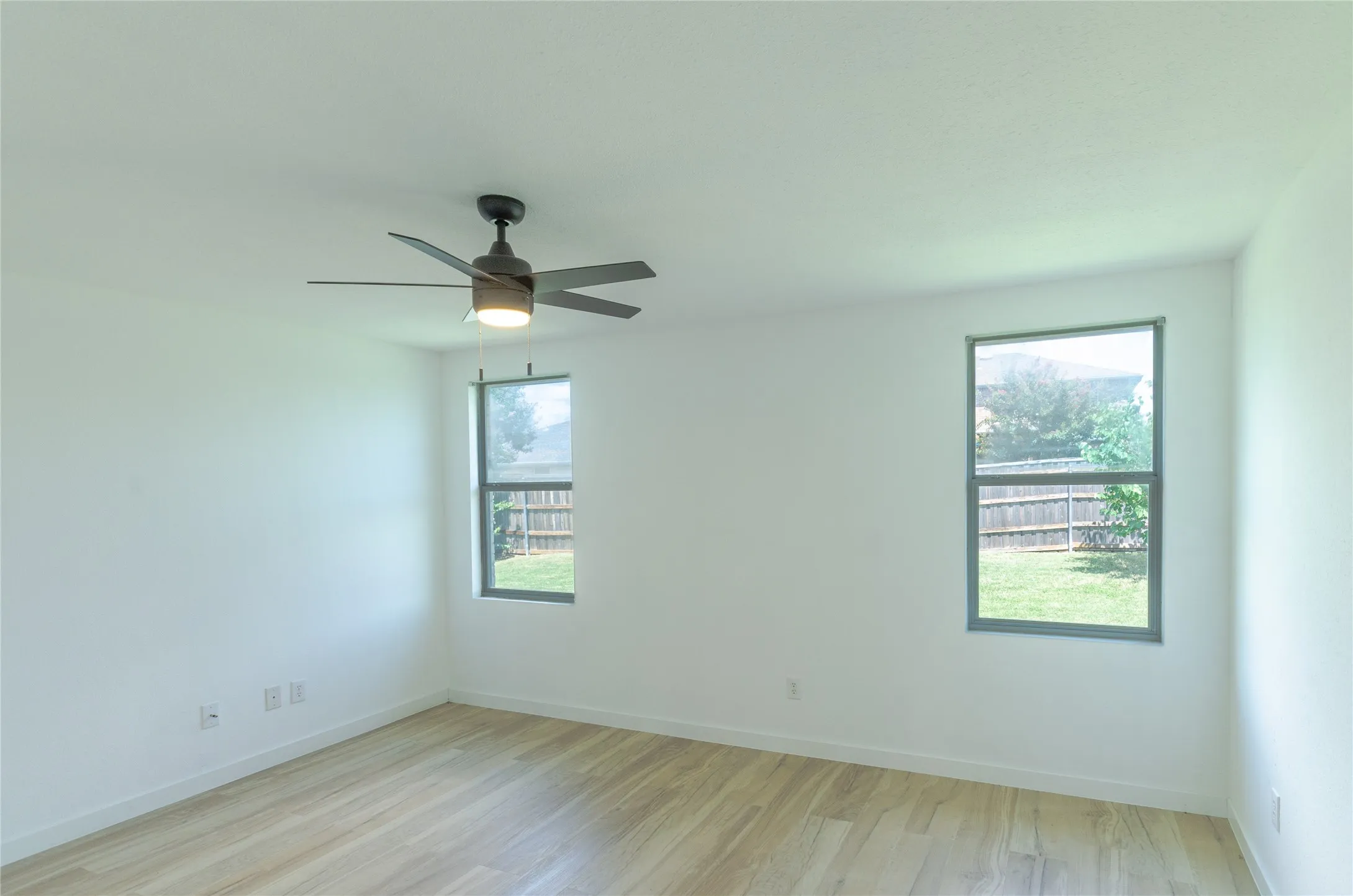 Bright room featuring wood-finish flooring, two windows, a ceiling fan with integrated lighting, and white walls