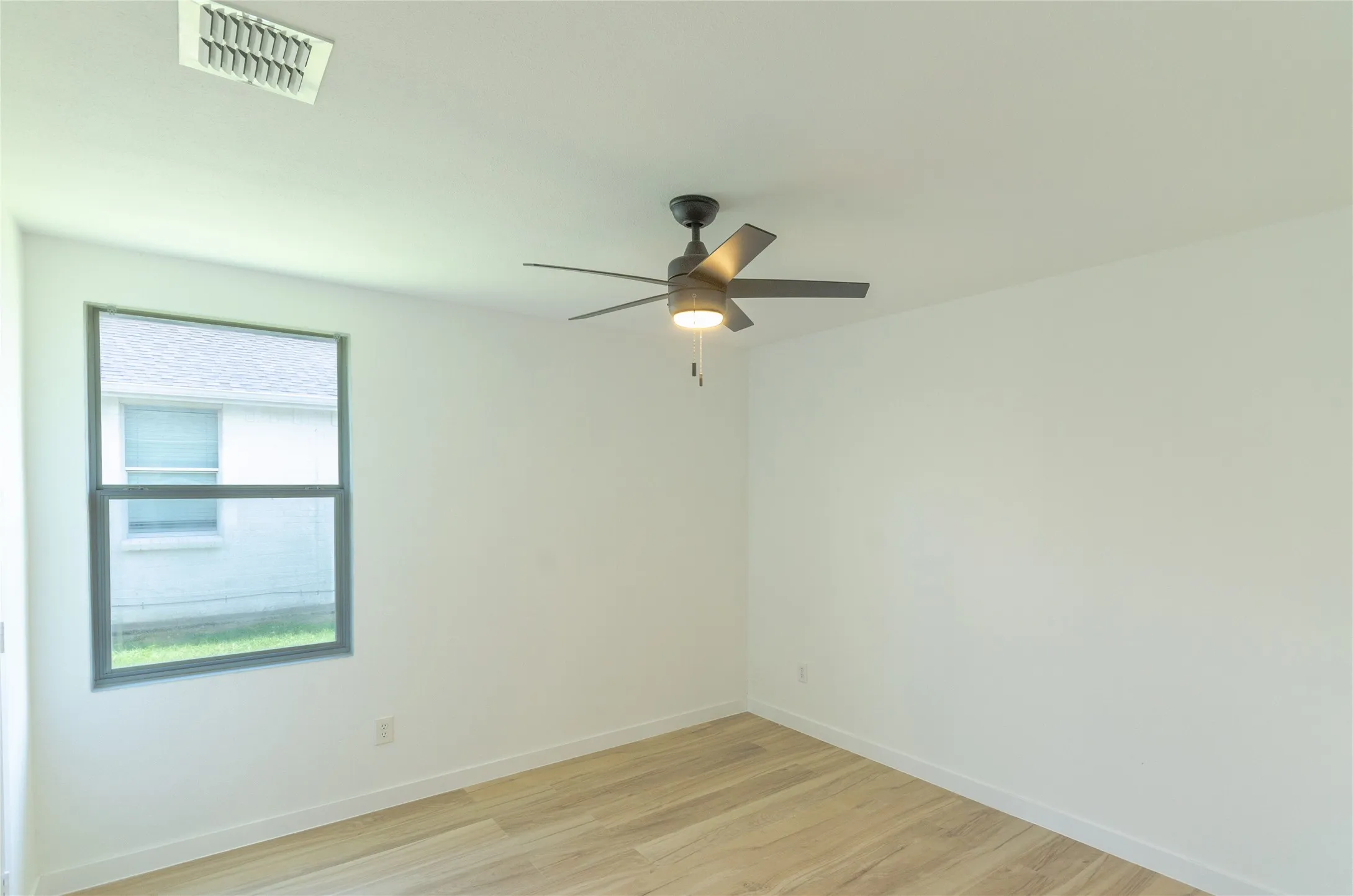Bright room featuring light wood-finish flooring, a modern black ceiling fan with integrated lighting, and a large window with grey trim