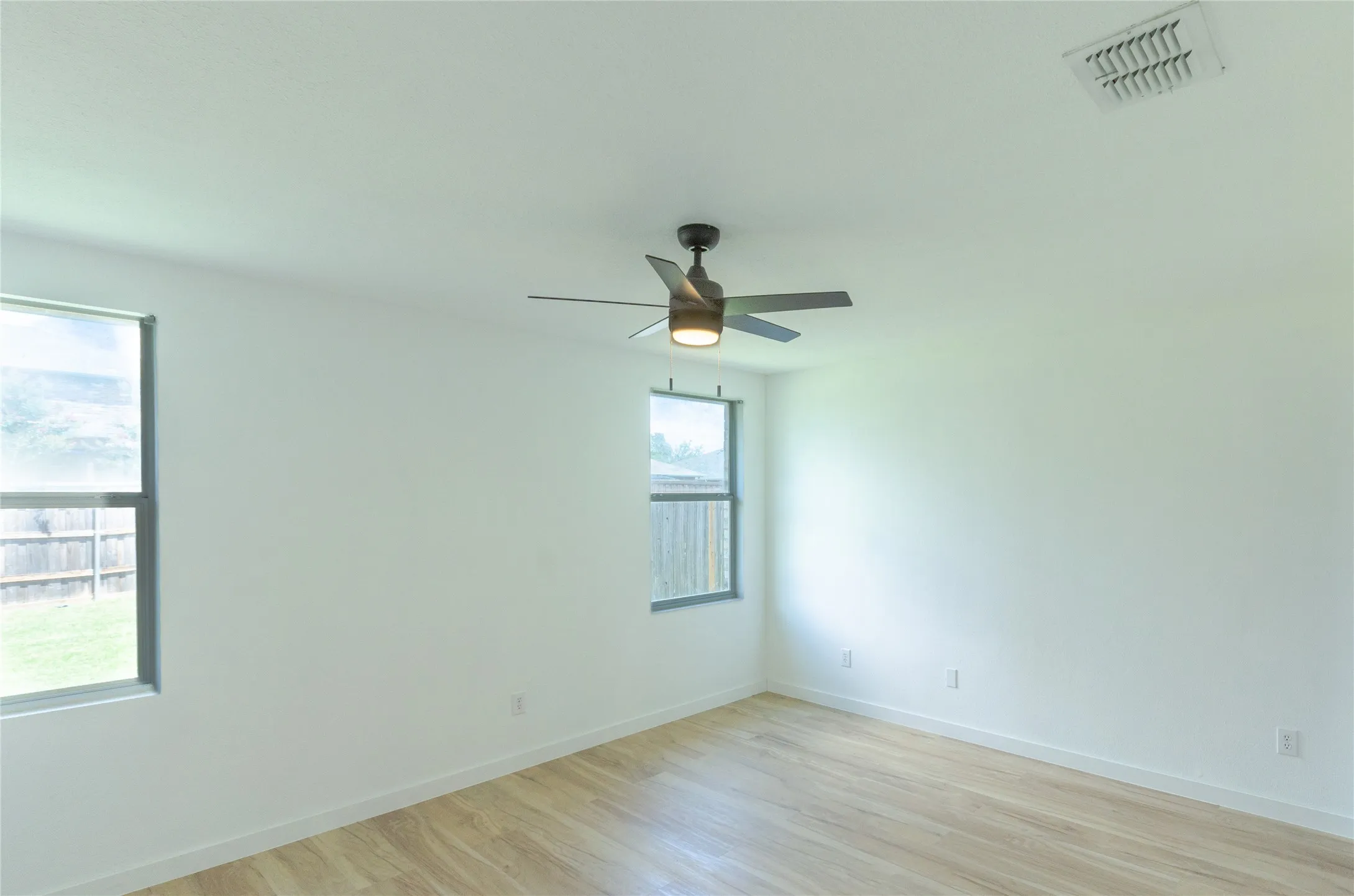 Bright interior space featuring light wood-finish flooring, two windows, a ceiling fan with integrated lighting, and white baseboards