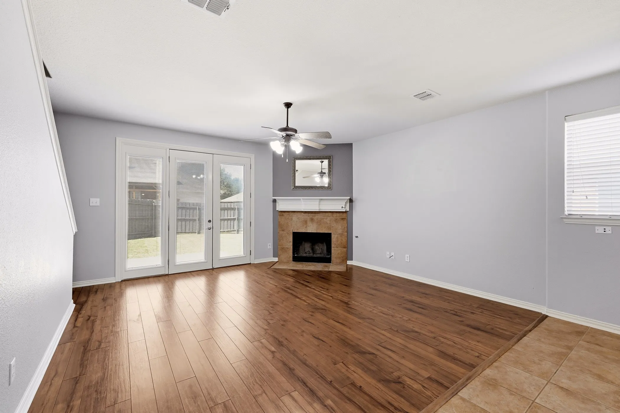 Spacious living area featuring rich wood-finish flooring, a corner fireplace with a stone surround and mantel, and triple glass-paned French doors