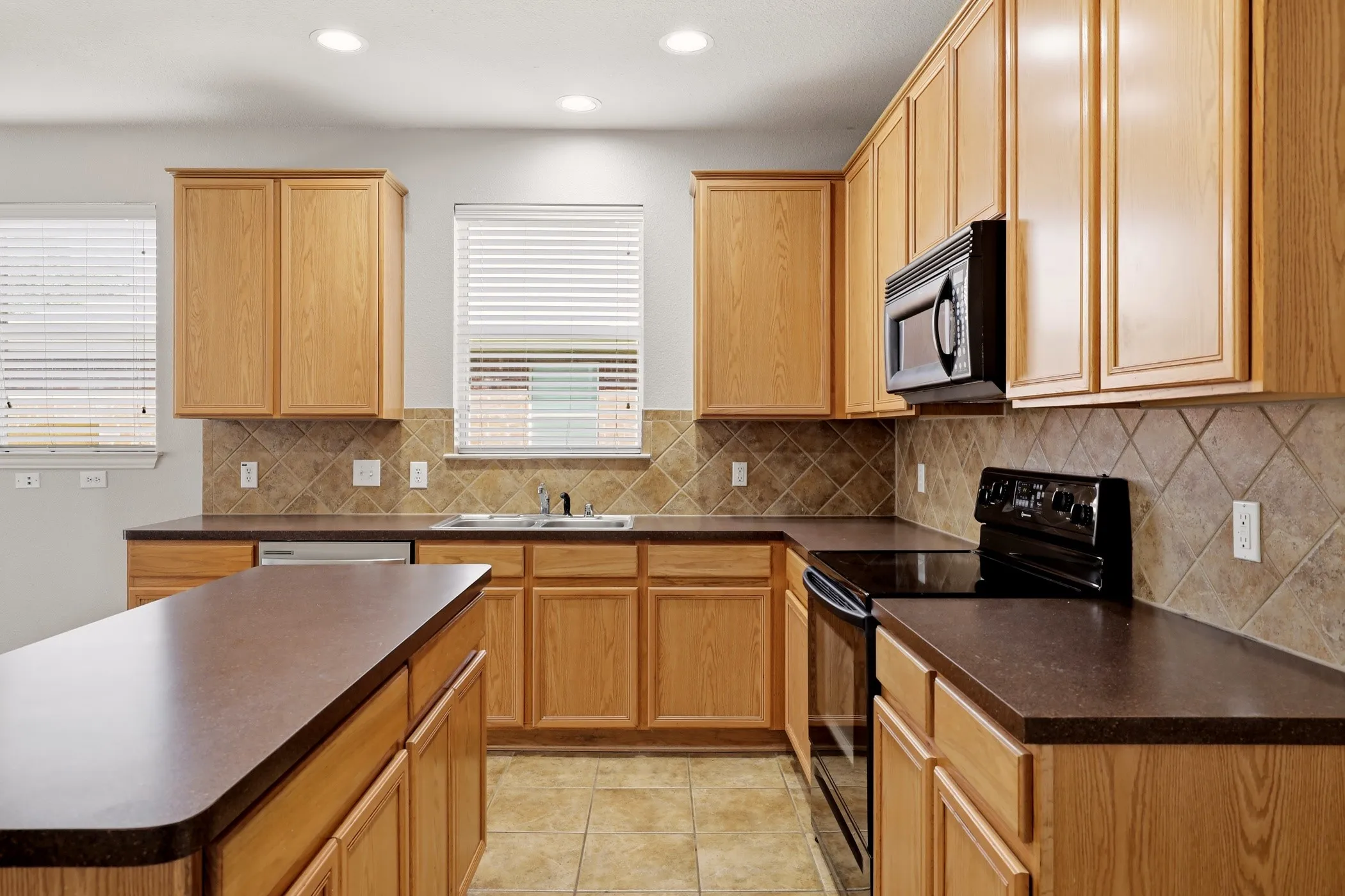 Kitchen featuring a central island, light wood cabinetry, dark countertops, a tile backsplash, and stainless-steel appliances including a built-in microwave and range