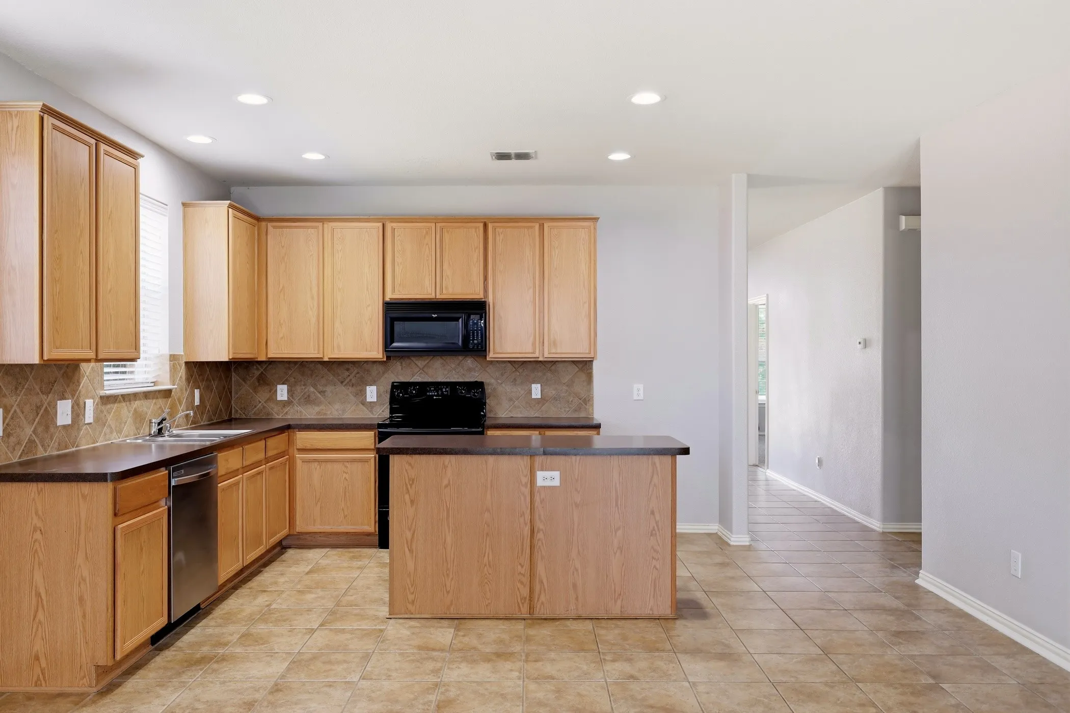 Kitchen featuring light wood cabinetry, dark countertops, a tiled backsplash, stainless steel dishwasher, and black appliances