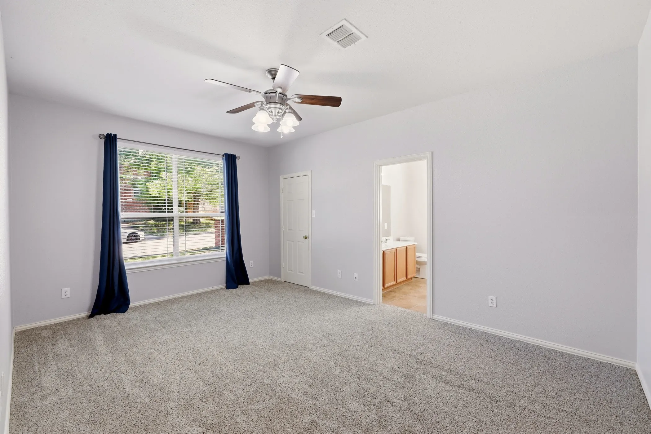 Spacious room featuring neutral carpet flooring, a ceiling fan with integrated lighting, a large window with blinds, and an open doorway leading to a bathroom with a wood-finish vanity