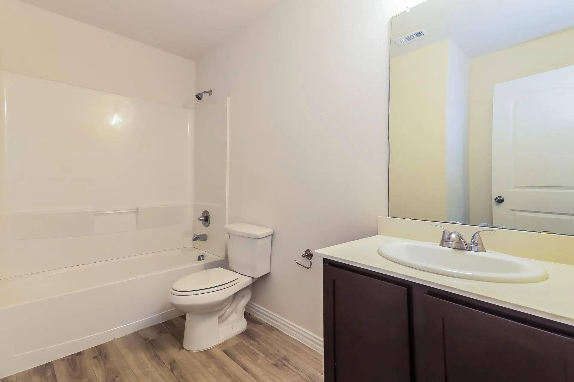 Bathroom featuring wood-finish flooring, a white fiberglass tub/shower combination, a white toilet, a vanity with dark wood-finish cabinetry, and a single basin sink with a chrome faucet