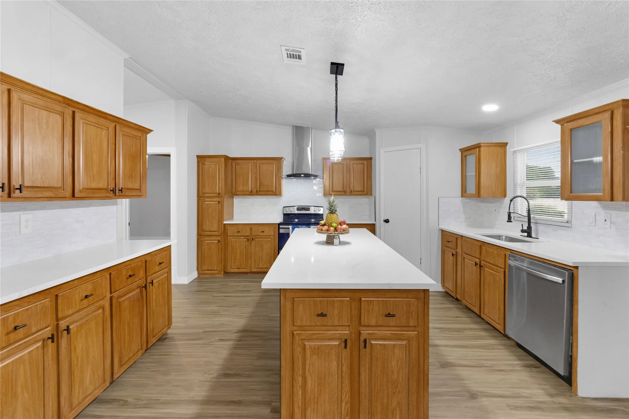 Open-concept kitchen featuring an island with a pendant light fixture, wood-finish cabinetry, light countertops, subway tile backsplash, and stainless steel appliances