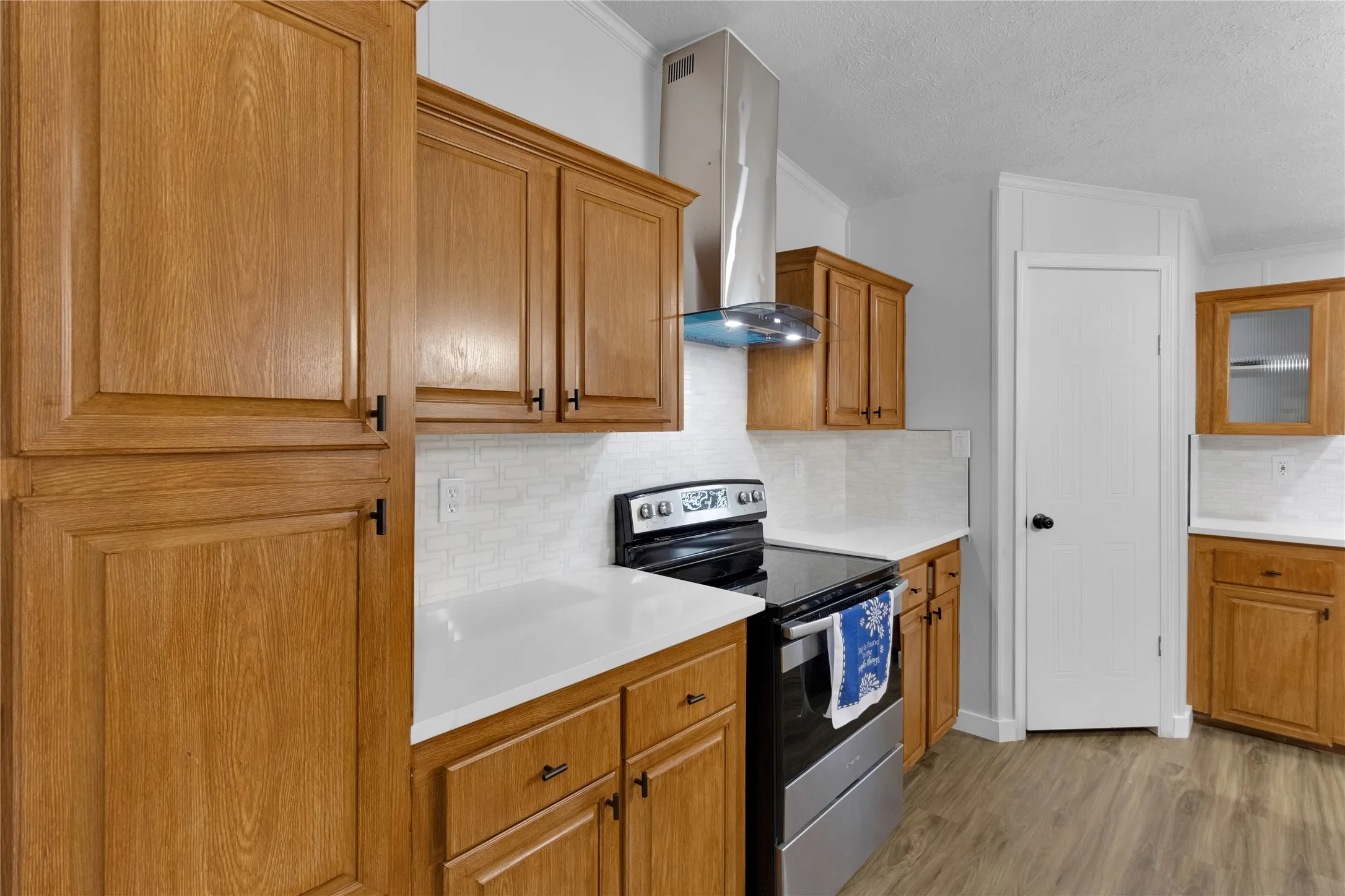 Kitchen featuring wood cabinetry, white countertops, a white subway tile backsplash, a stainless steel range hood, and wood-finish flooring