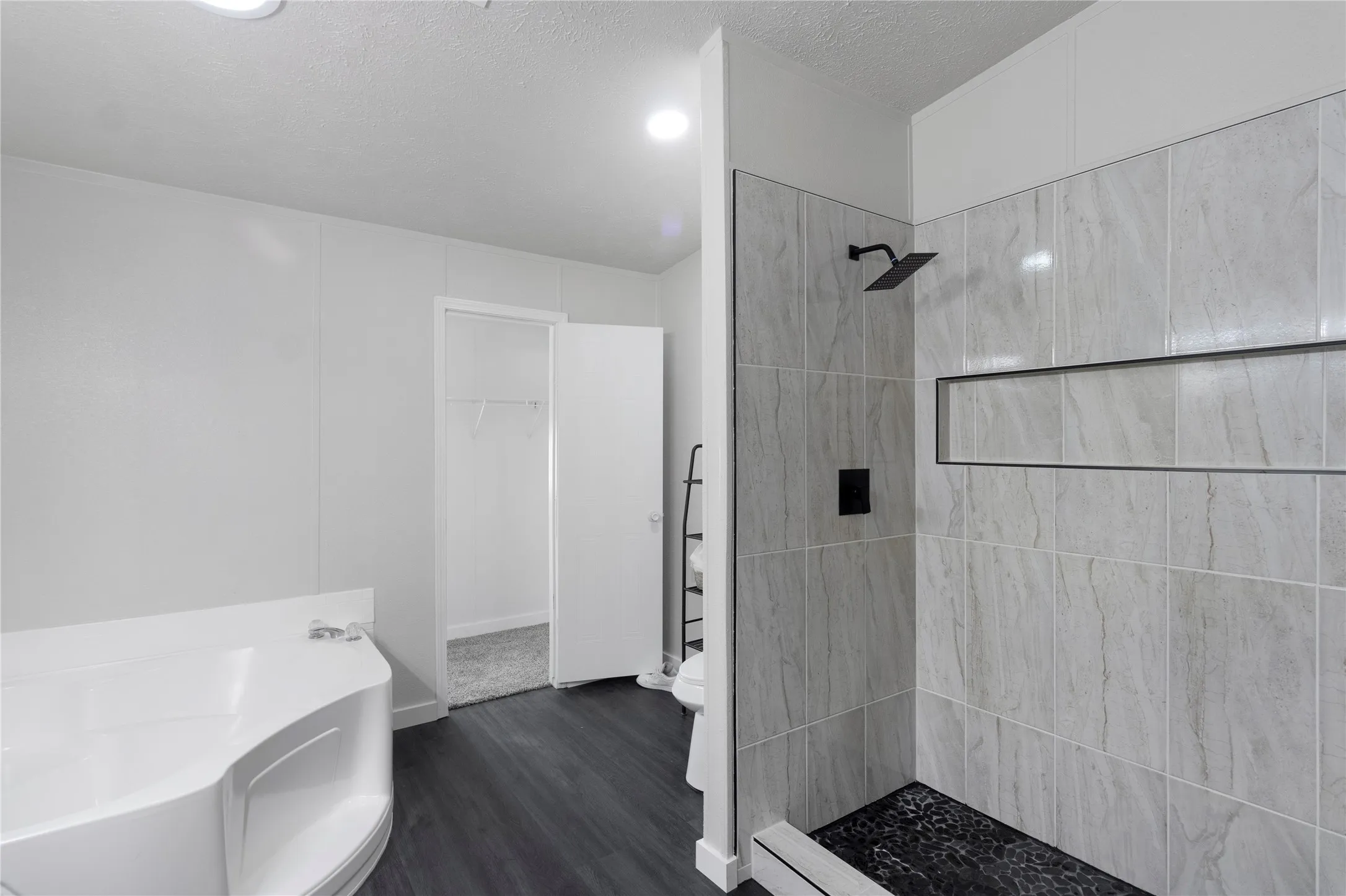 Spacious bathroom featuring a built-in soaking tub, a walk-in shower with light-toned tile walls and black pebble flooring, dark wood-finish flooring, and recessed lighting
