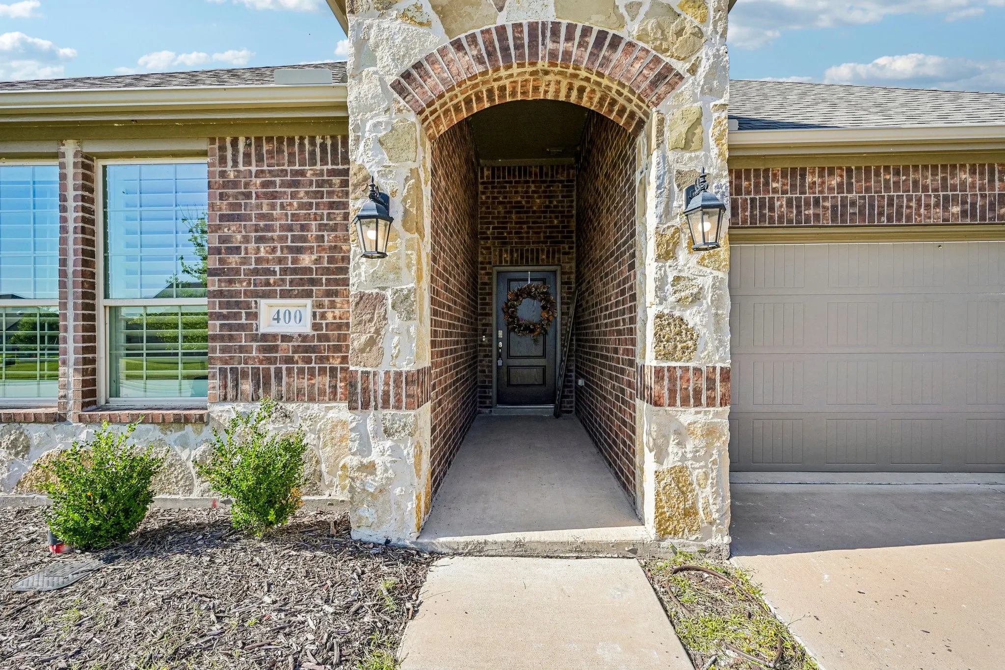 Brick and stone exterior featuring an arched entryway, two decorative exterior sconces, a paneled garage door, and a concrete pathway