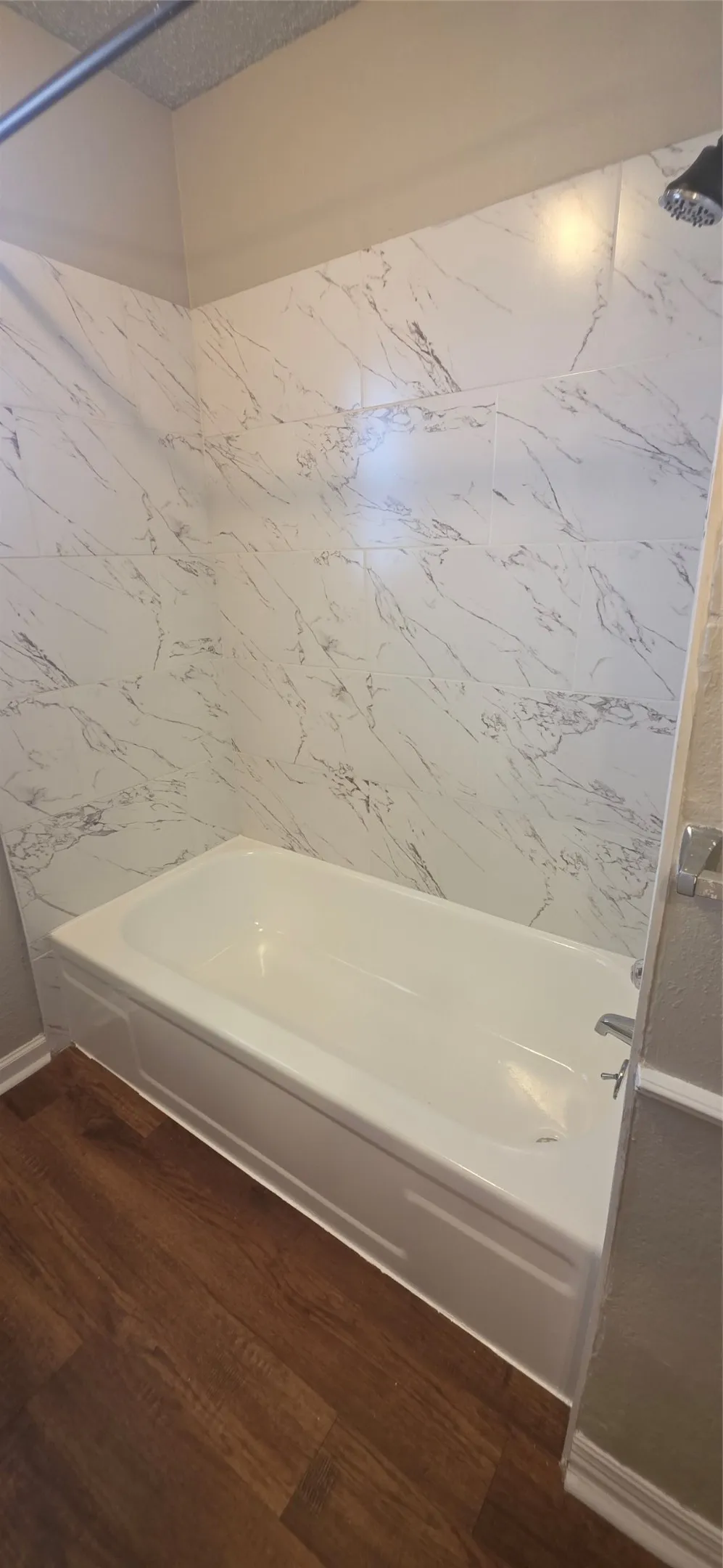 White soaking tub with a wall-mounted showerhead, surrounded by white tile with grey veining, and wood-finish flooring