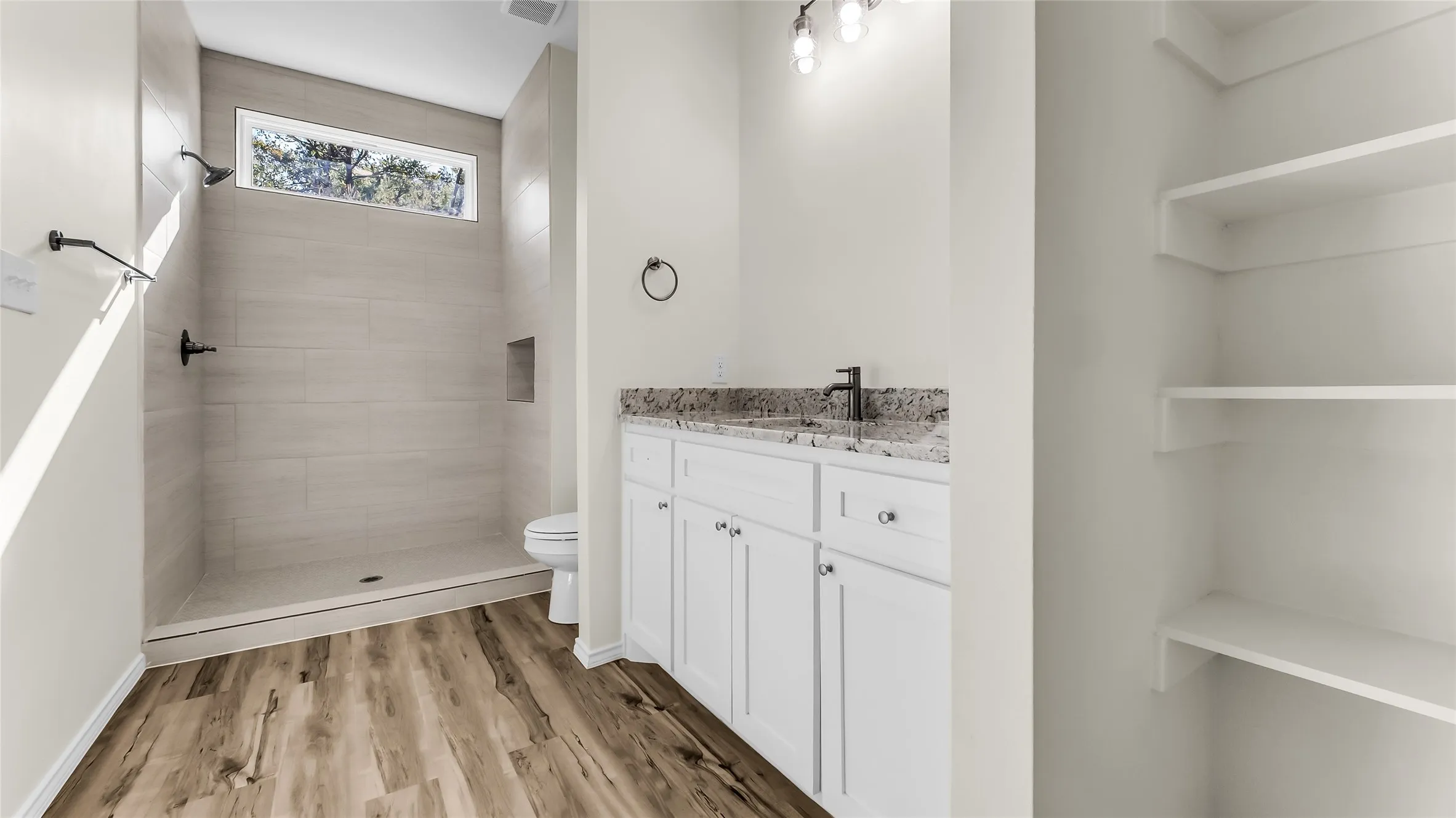 Walk-in shower with tile surround, built-in shelving, wood-finish flooring, vanity with stone countertop, and integrated shelving