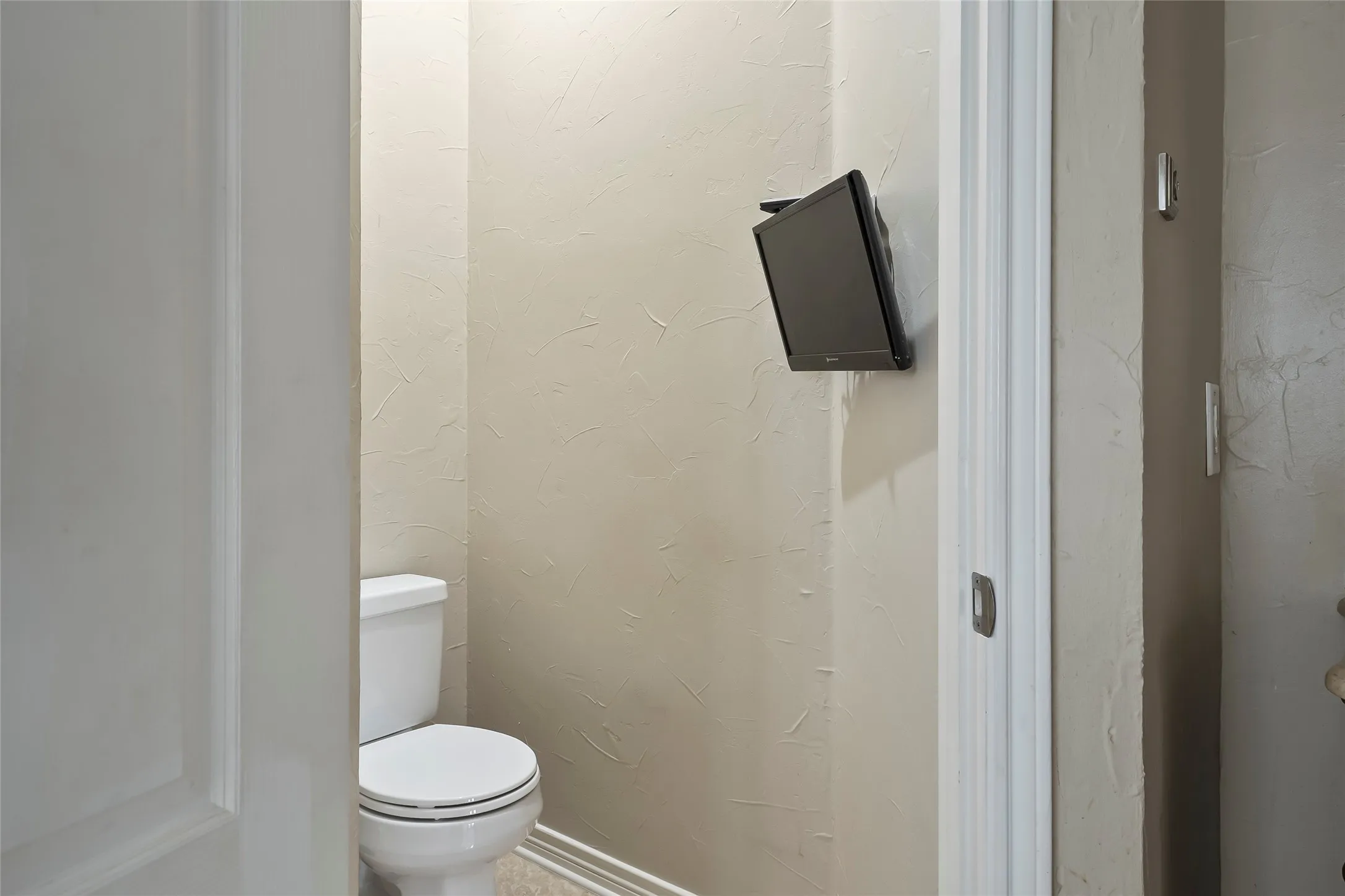 White toilet with a built-in wall-mounted television, textured beige wall finish, and white baseboard molding