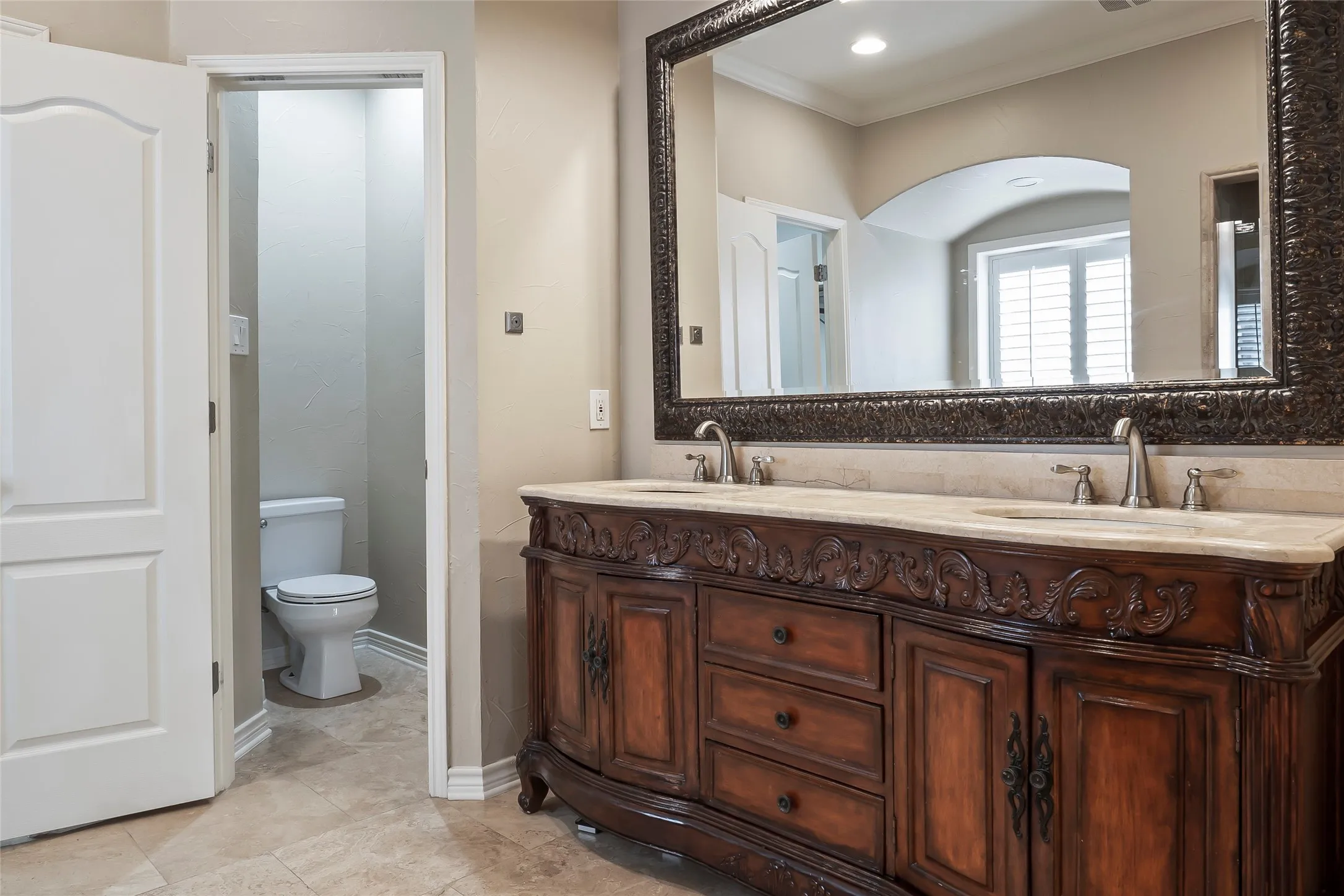 Dual sink vanity with carved wood cabinetry, stone countertop, and ornate framed mirror