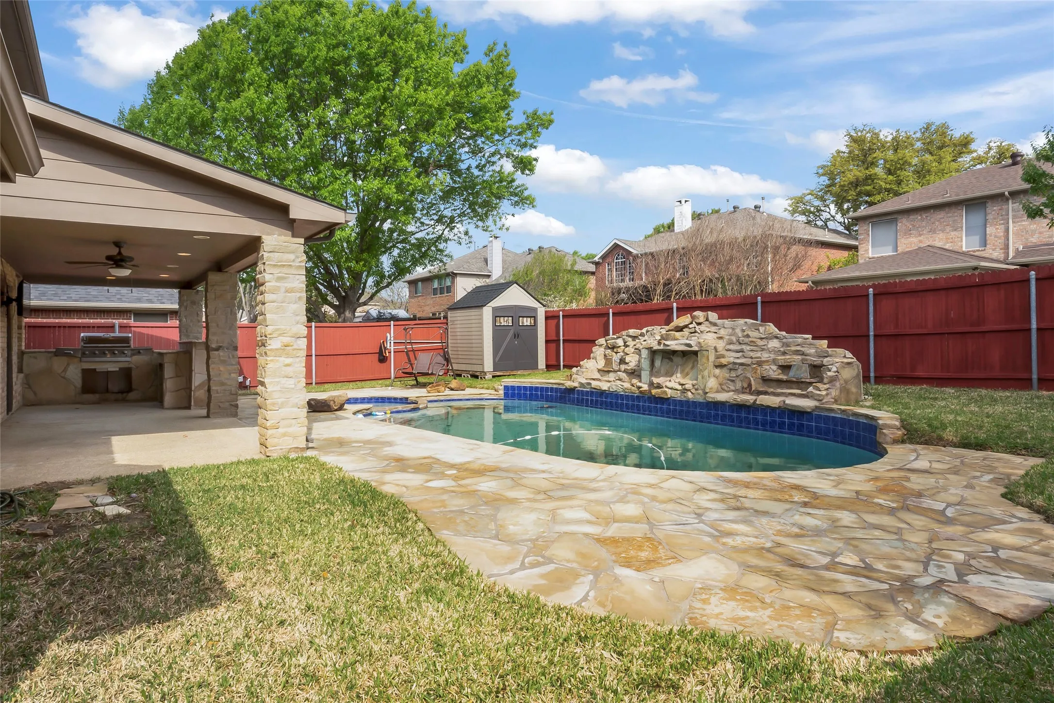 Backyard featuring a freeform swimming pool with a rock waterfall, a covered patio with an outdoor kitchen, flagstone decking, a storage shed, and a privacy fence
