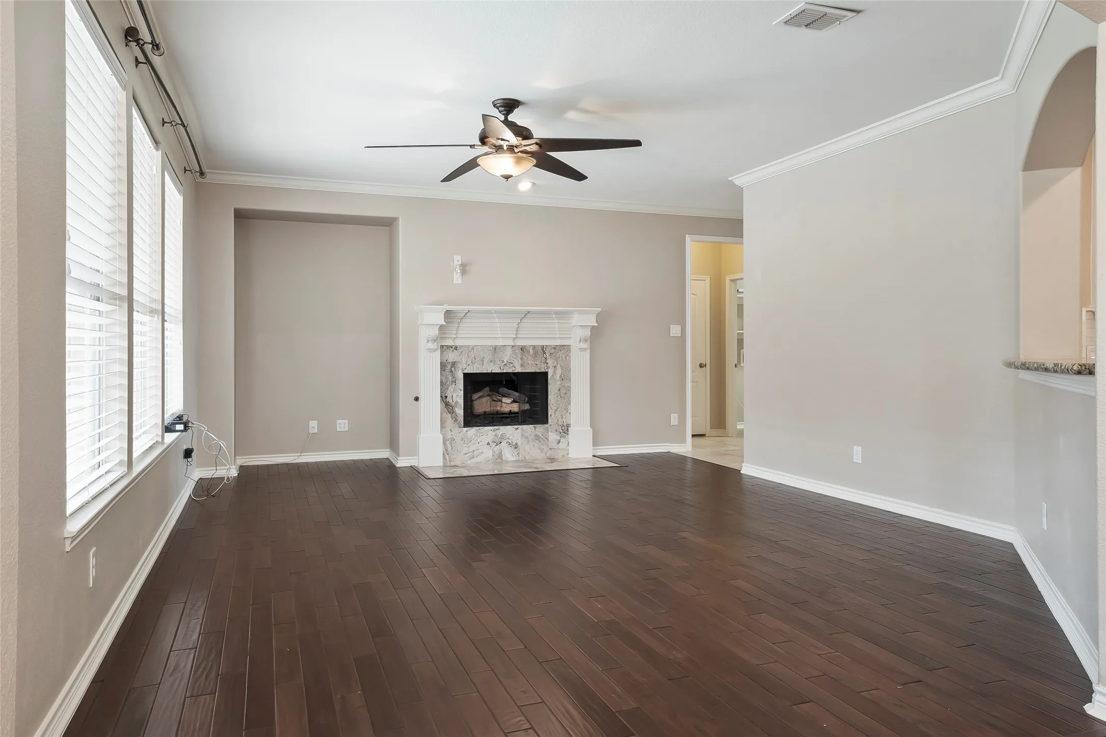 Spacious living area featuring dark wood-finish flooring, a white mantel fireplace with a stone surround, multiple windows with blinds, a ceiling fan, and crown molding