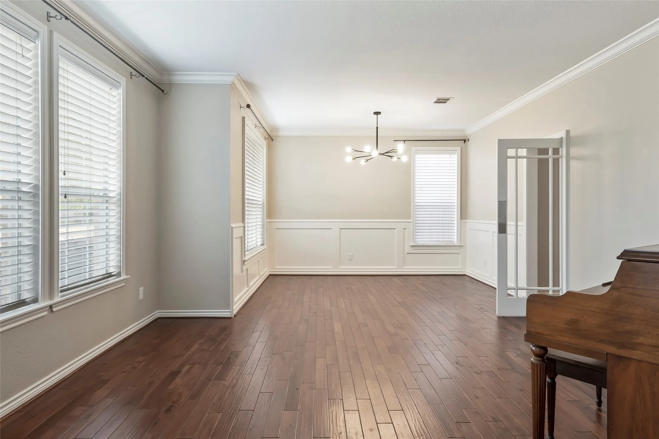 Spacious room featuring wood-finish flooring, white paneling, multiple windows with blinds, and a modern chandelier