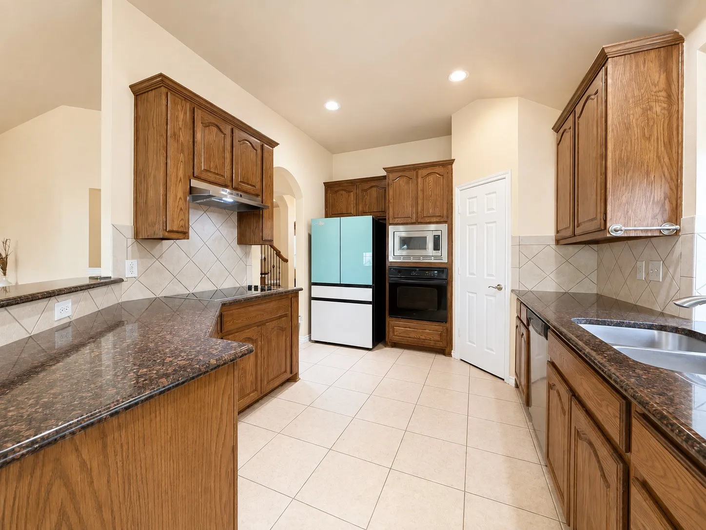 Galley-style kitchen featuring warm wood cabinetry, granite countertops, diagonal tile backsplash, stainless steel appliances including a microwave and double wall ovens, and ceramic tile flooring throughout