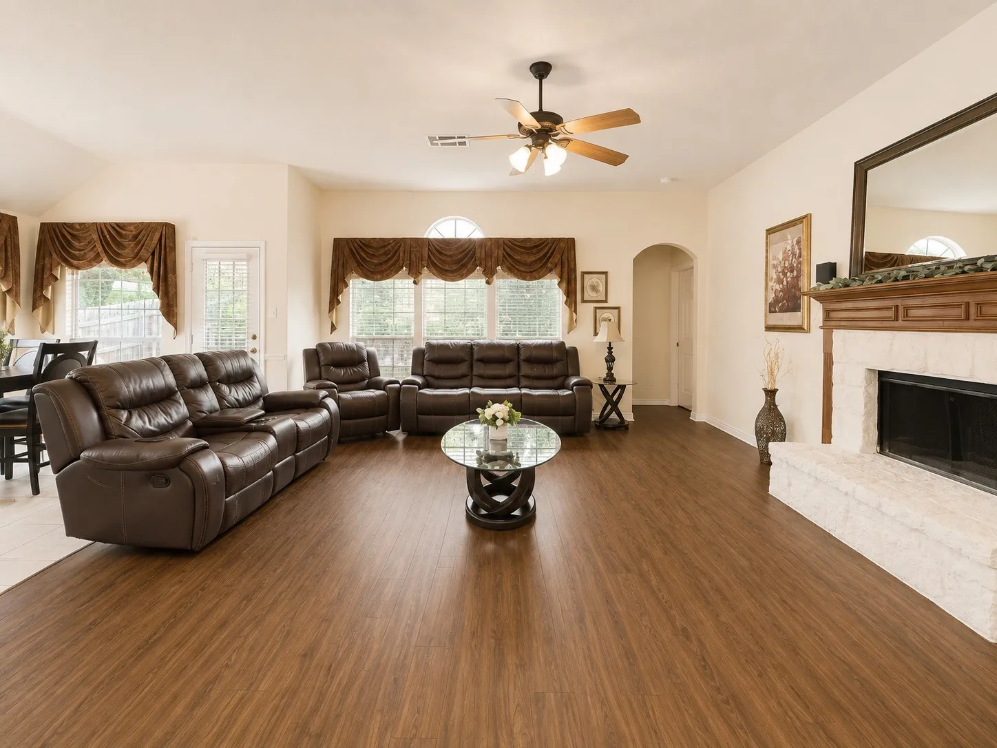 Spacious living room featuring warm wood-finish flooring, a ceiling fan with integrated lighting, and a fireplace with a stone surround and wooden mantel