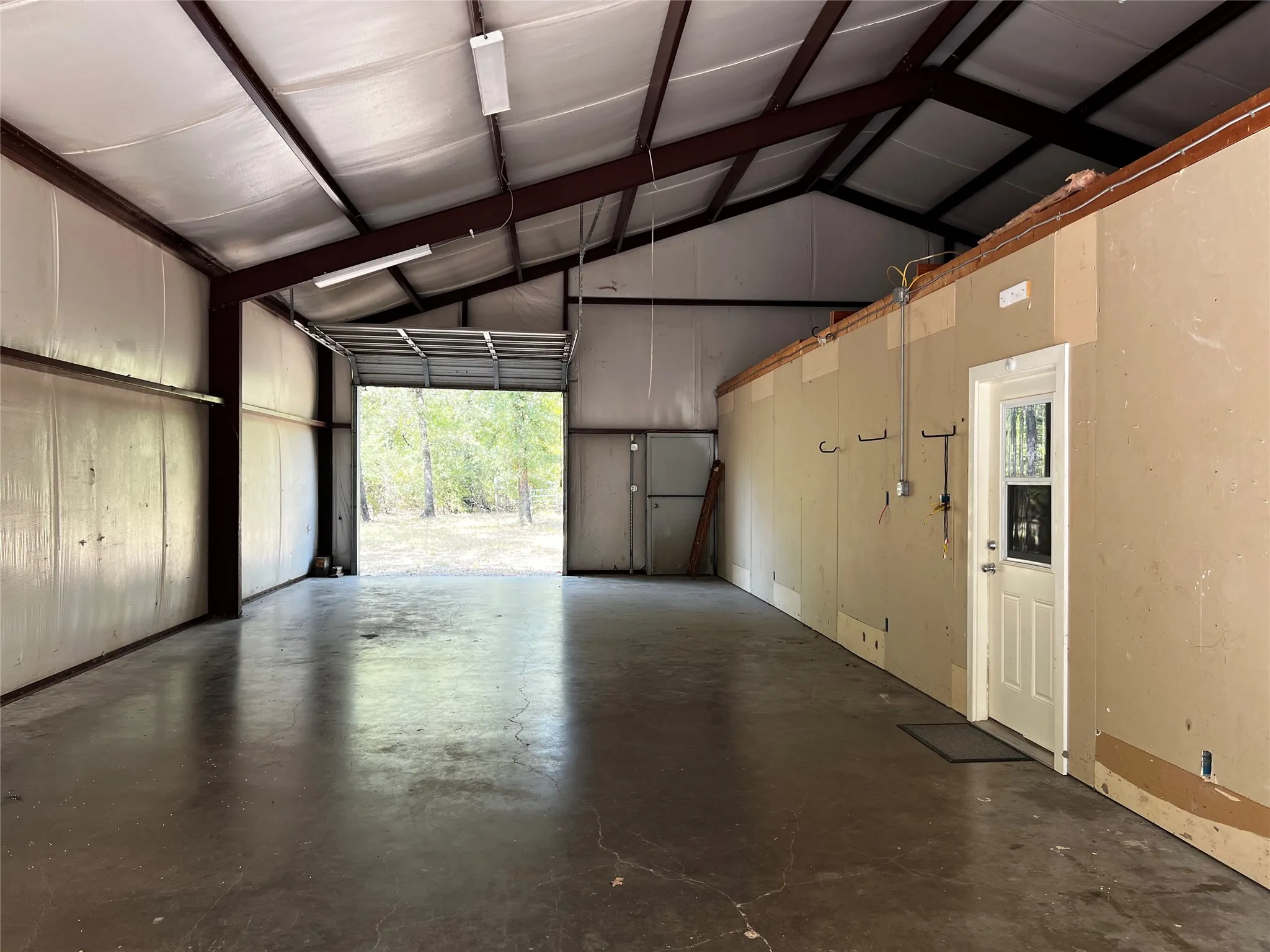 Garage - Workshop - Expansive open interior with concrete flooring, insulation, exposed structural beams, 12' eave height and 10' high roll-up single garage door and exterior entry door at north end of garage.  Looking from from south end roll-up garage door.  Note entry door on wall to the Barndo Living Space.