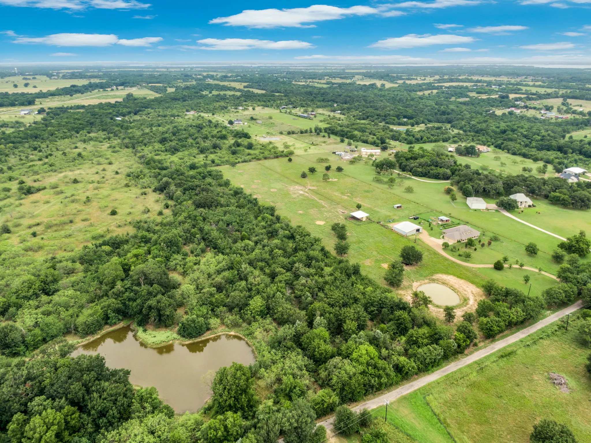 Expansive rural landscape featuring a private pond, abundant mature trees, open pastureland, and scattered residential properties