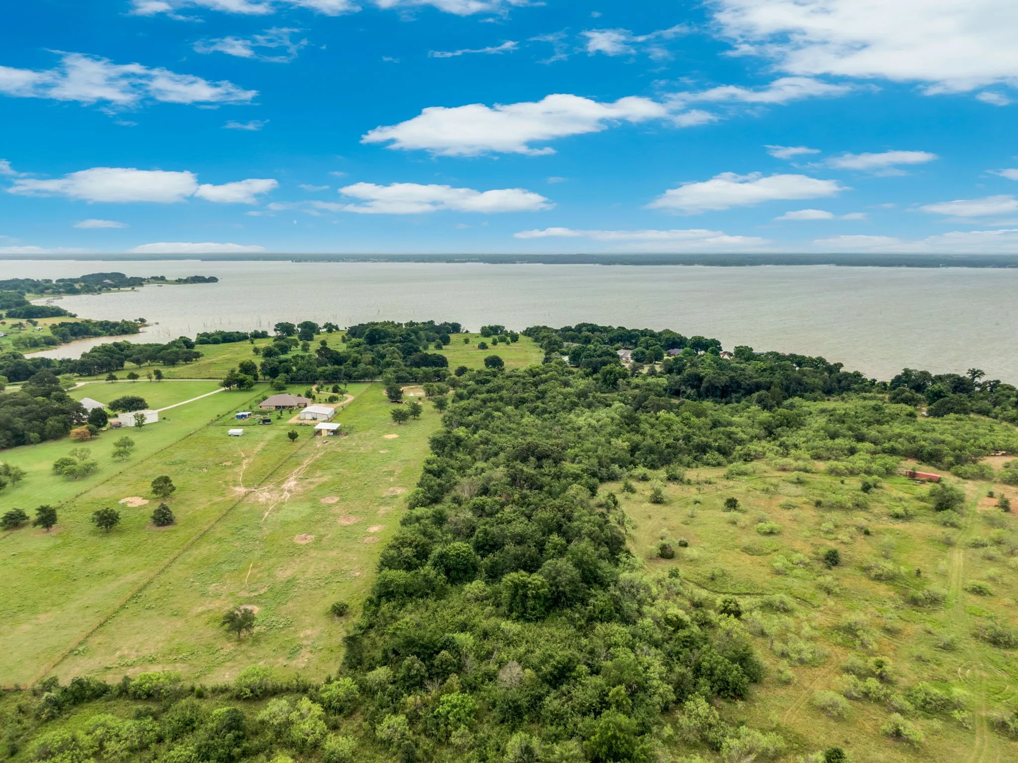 Expansive waterfront property featuring a large lake, verdant fields, a mix of mature trees and brush, and a visible residence with outbuildings