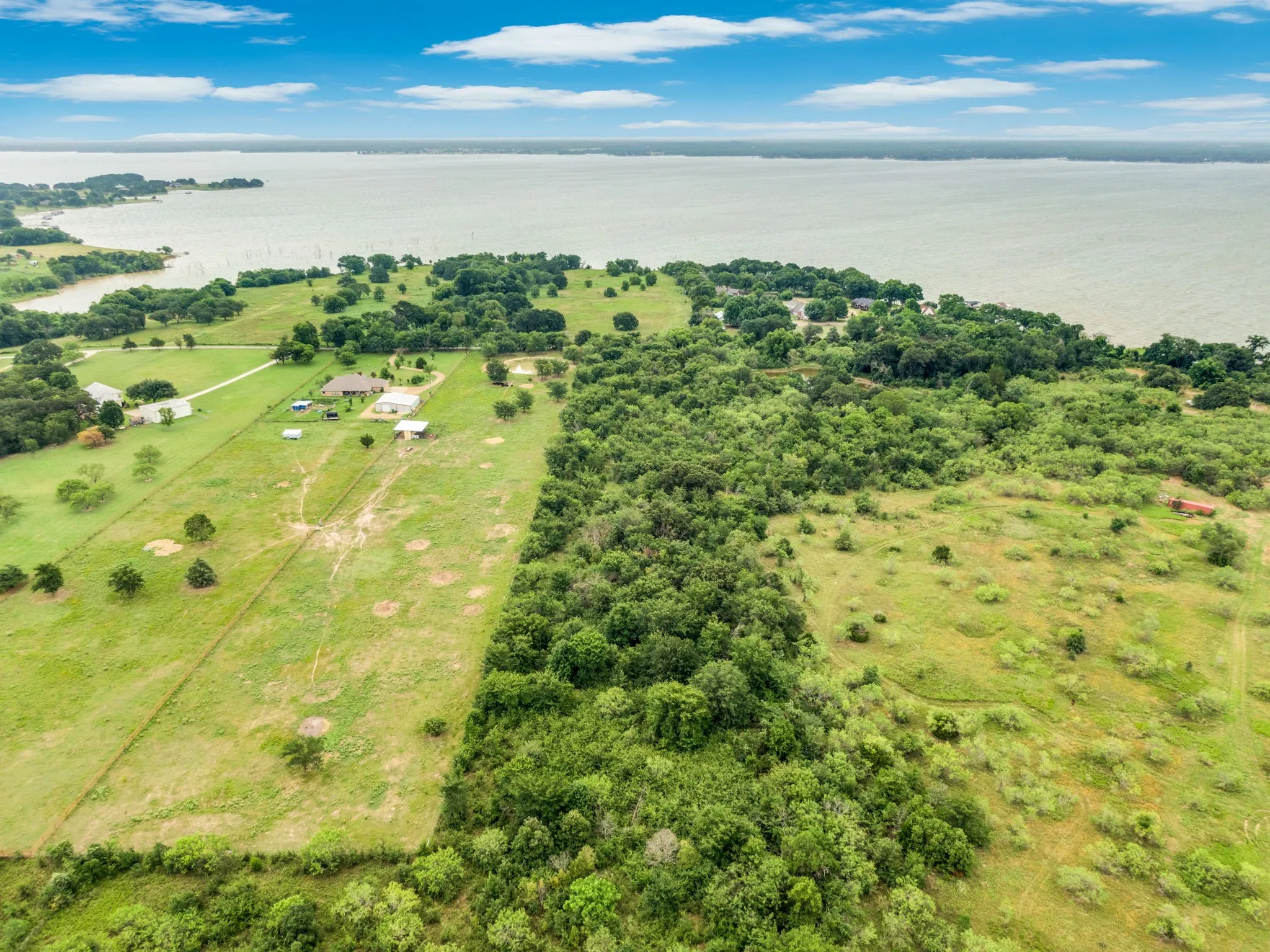 Expansive waterfront property featuring a large body of water, extensive green acreage, substantial tree coverage, and multiple structures