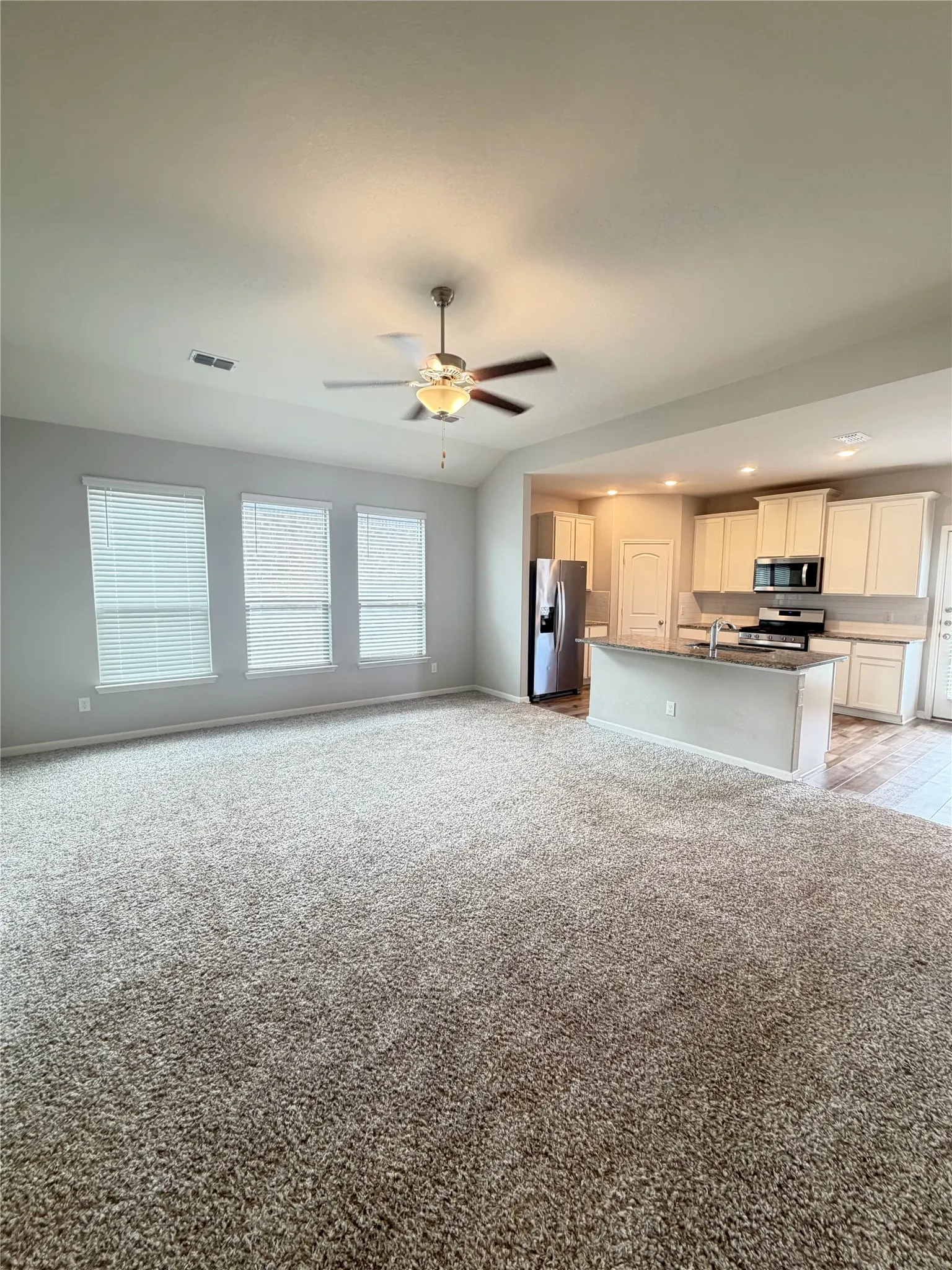 Spacious living area featuring neutral-toned carpeting, a ceiling fan, and three windows with horizontal blinds