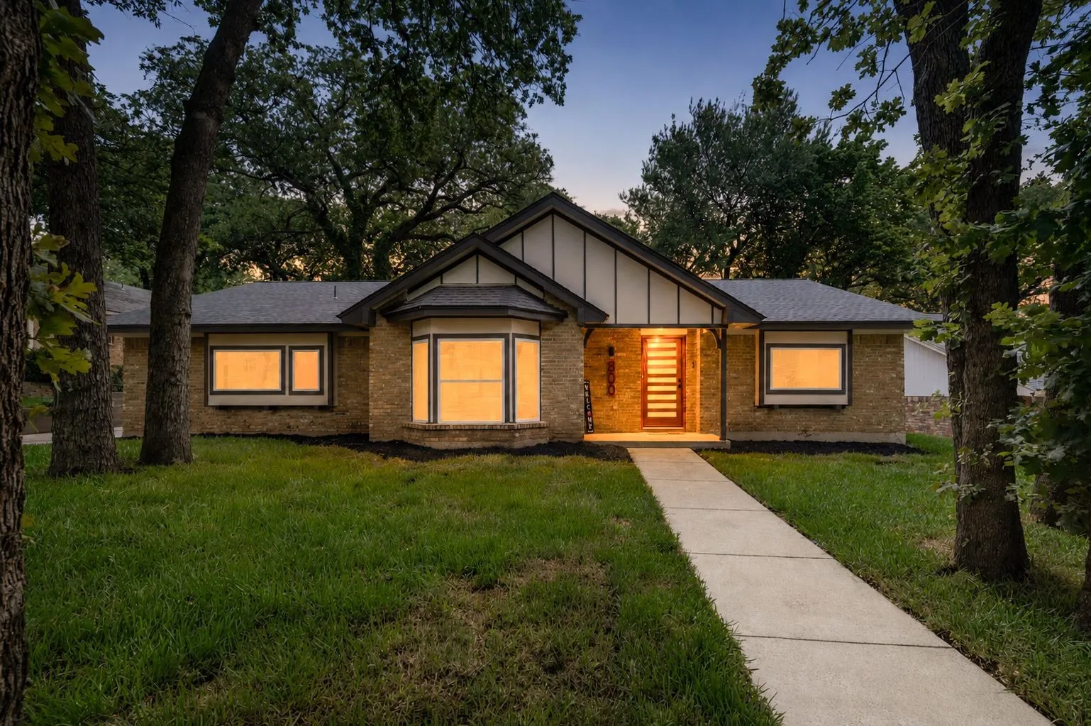 As evening settles in, warm interior light turns the home into an inviting retreat beneath the trees.