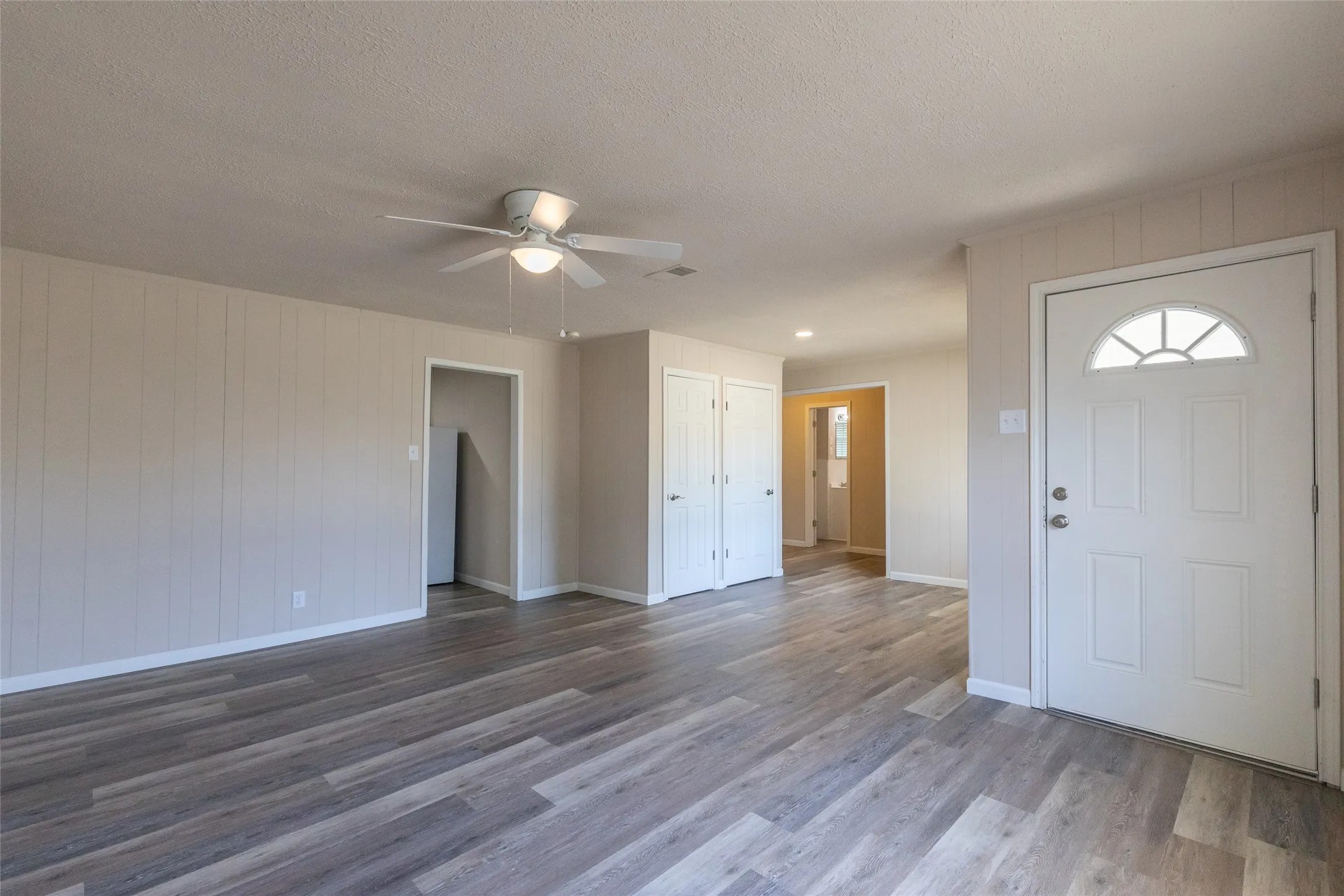 Spacious room featuring wood-finish flooring, light-toned walls with vertical paneling, and a ceiling fan with integrated lighting