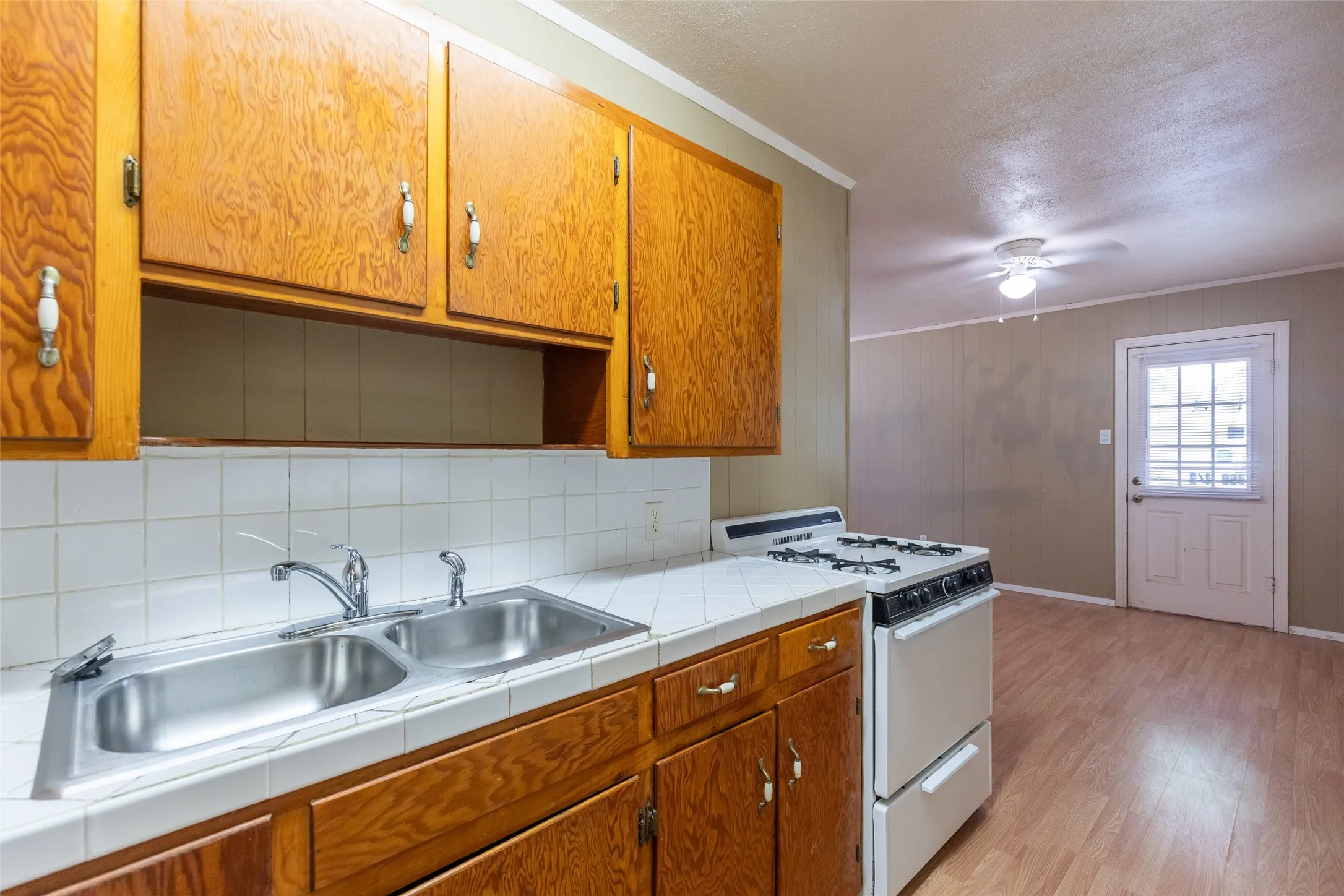 Kitchen featuring wood-finish cabinetry, a double basin stainless steel sink, white tile countertops, and a white gas range