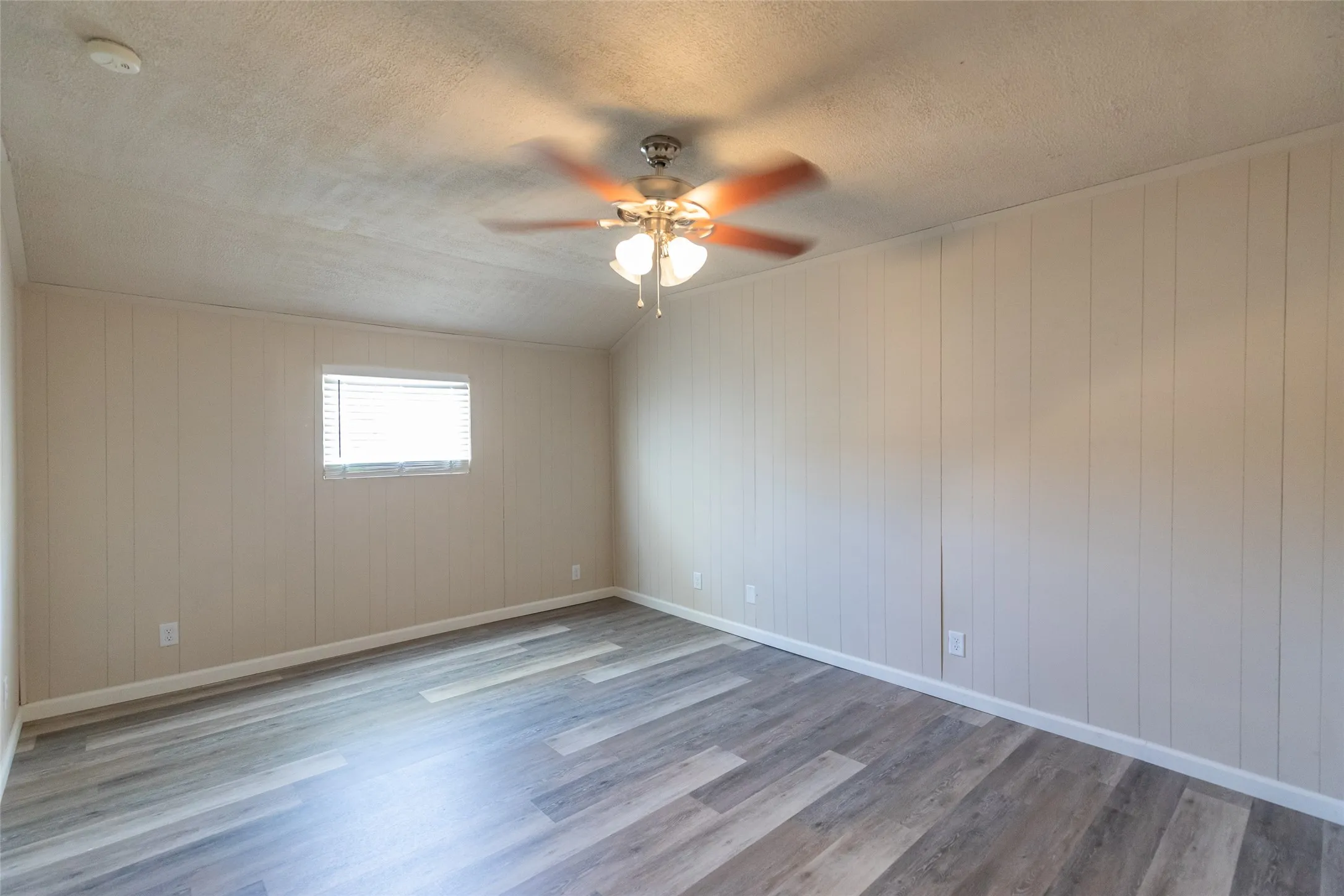 Room featuring wood-finish flooring, vertical paneling on walls, a ceiling fan with integrated lighting, and a single window with blinds