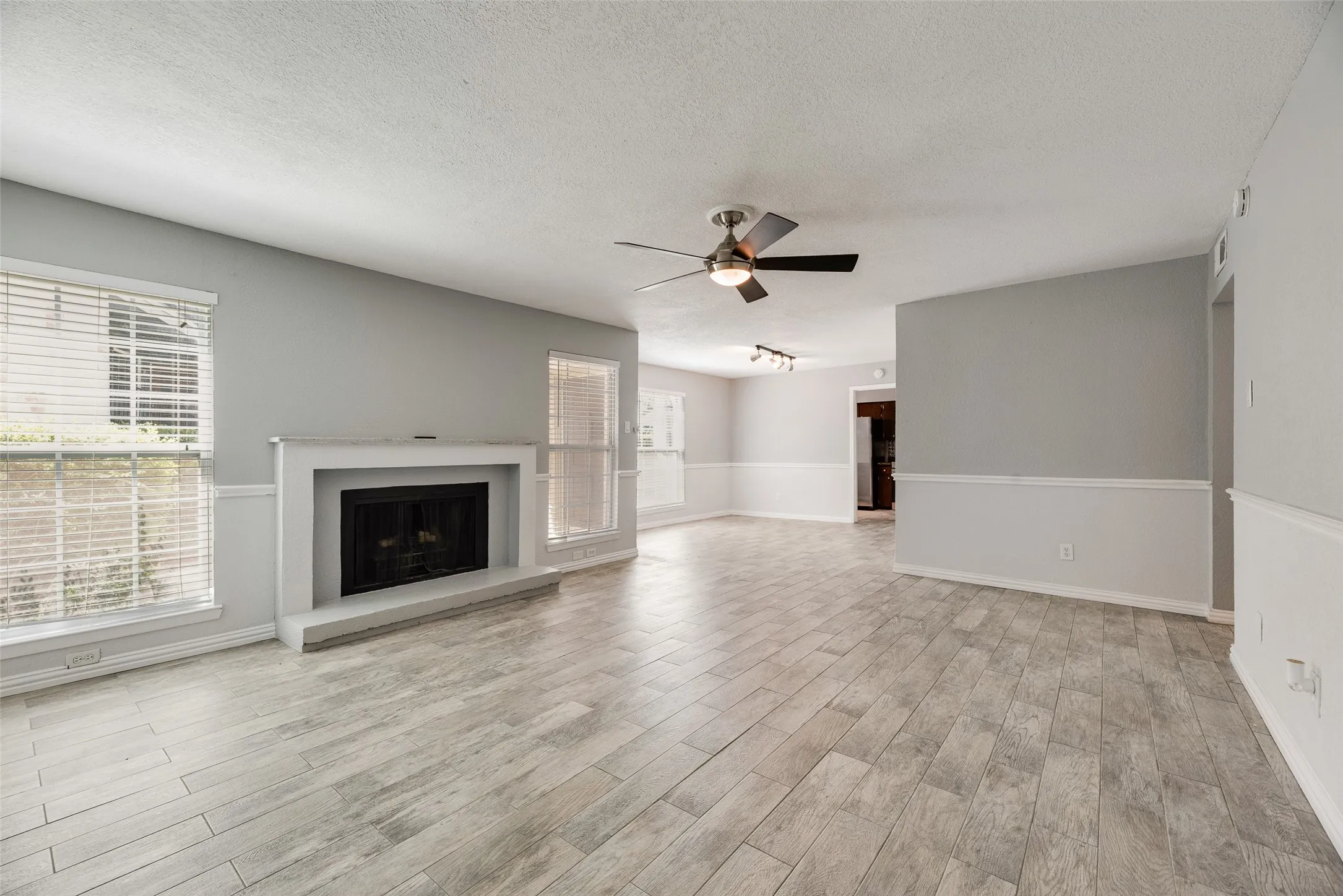 Spacious living area featuring a fireplace with a white mantel, light wood-finish flooring, and multiple windows with blinds