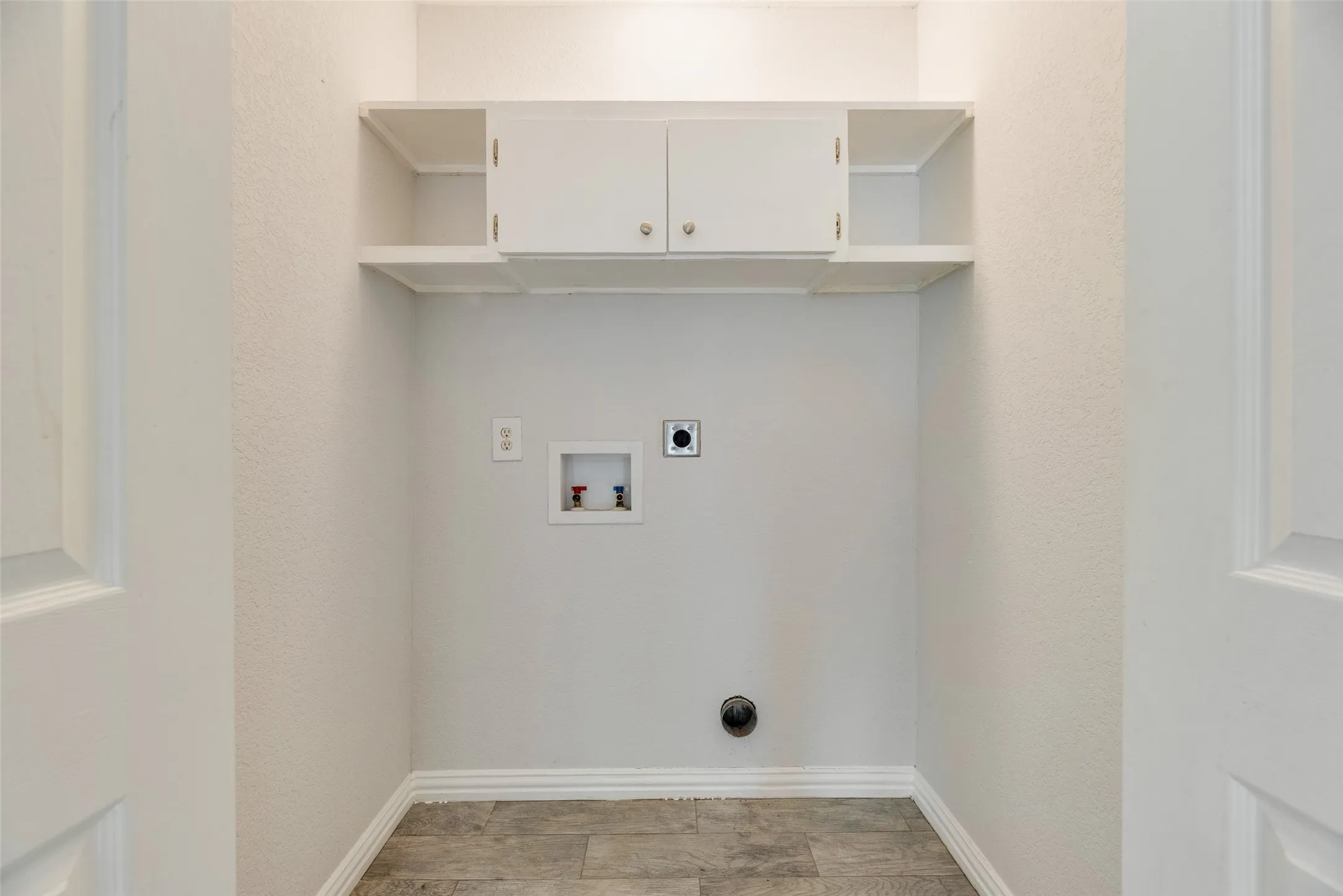 Dedicated laundry area featuring overhead white cabinetry with silver hardware, open shelving, and wood-finish flooring