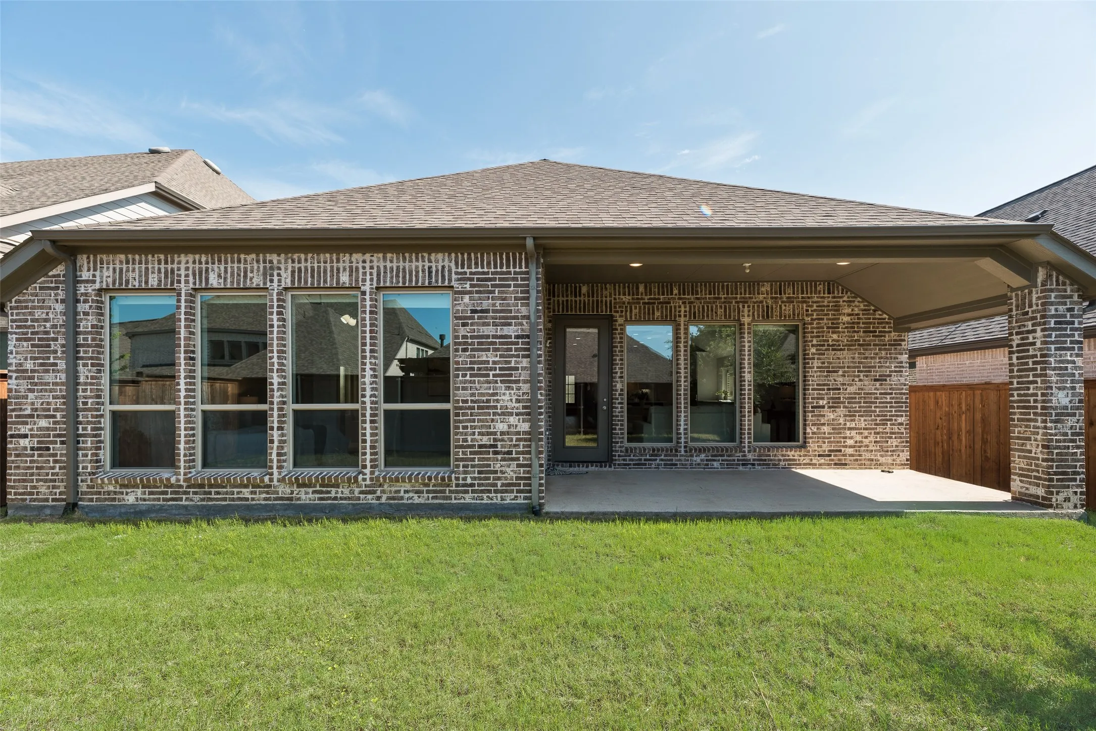 Expansive backyard featuring a manicured lawn, a full-brick exterior, multiple large windows, a covered patio with recessed lighting, and a private wood fence