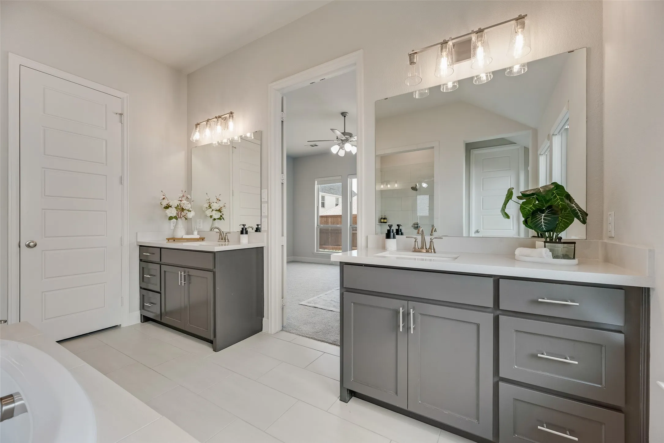 Dual vanity bathroom featuring gray cabinetry, white countertops, large mirrors with overhead lighting, and light-toned tile flooring