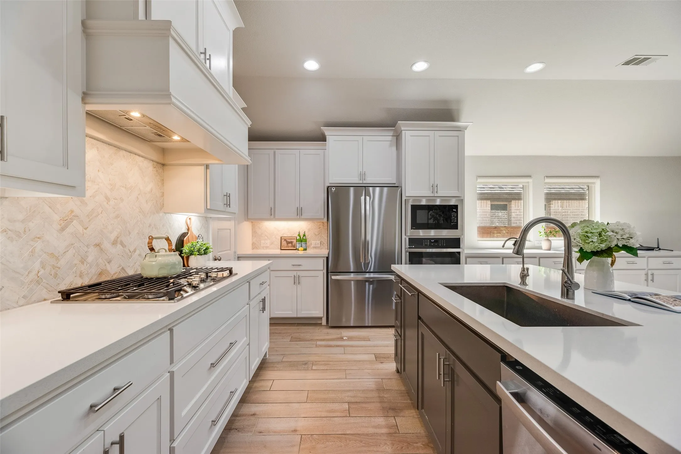 Gourmet kitchen featuring a built-in gas cooktop with decorative hood, herringbone pattern backsplash, white shaker cabinetry, stainless steel appliances, and a central island with an undermount sink