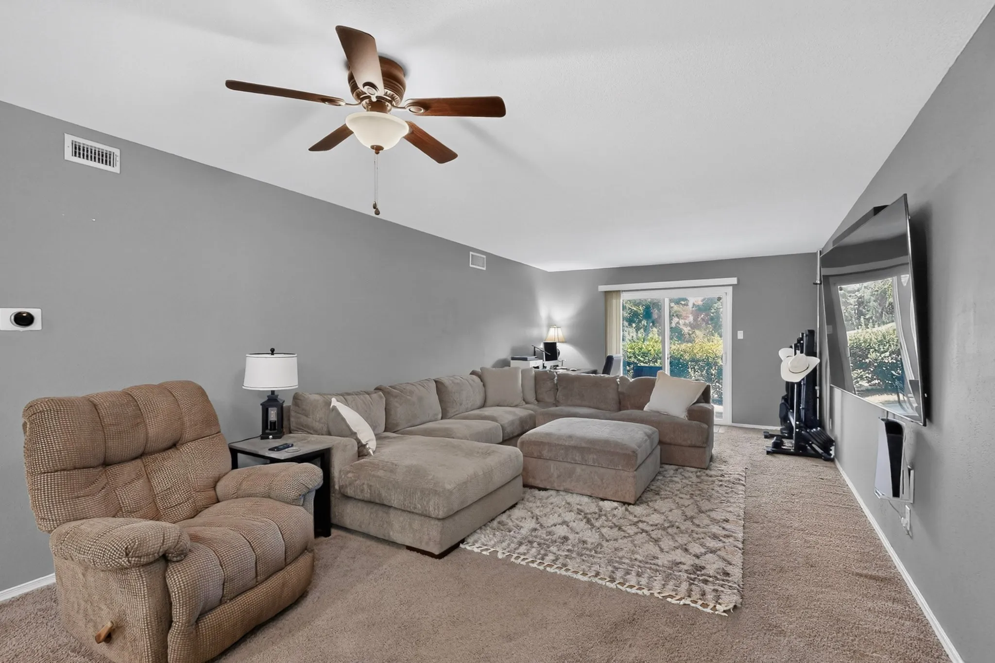 Living area featuring gray wall paint, a ceiling fan with integrated lighting, recessed wall vents, and sliding glass doors providing access to exterior greenspace