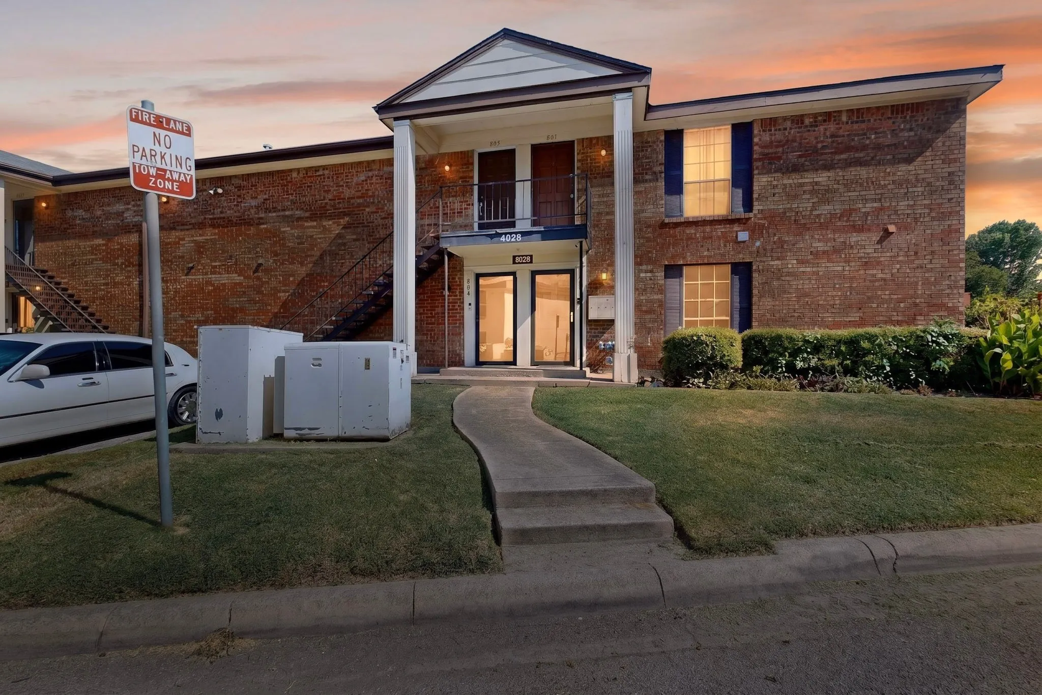 Brick exterior building featuring a covered entryway with white columns, a second-story balcony with metal railing, and dark window shutters