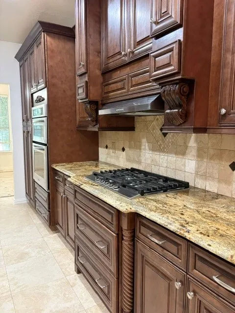 Rich wood cabinetry with decorative corbels and fluted accents, under-cabinet lighting, a gas cooktop, double wall ovens, and stone countertops